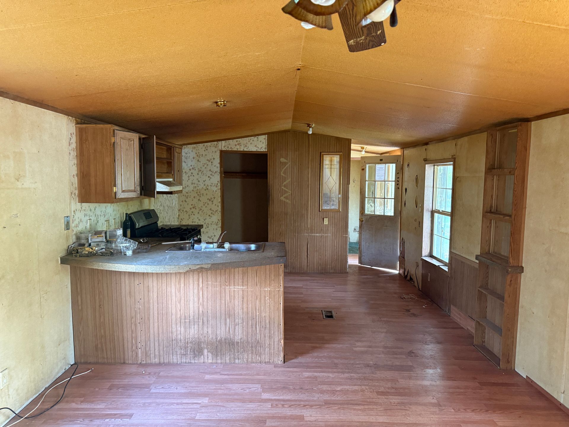 Interior of a mobile home kitchen and living area, with wood paneling, cabinets, and a low ceiling.