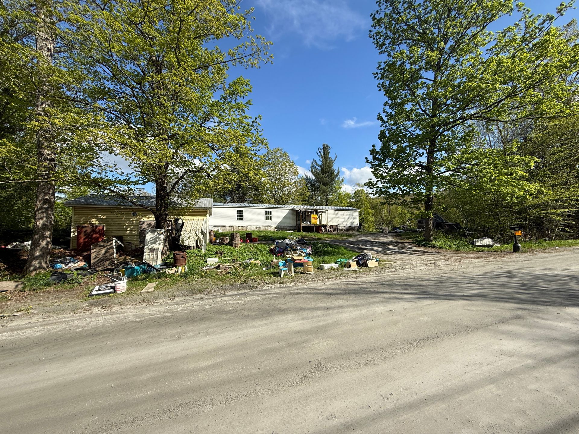 Dirt road leads to a mobile home surrounded by trees on a sunny day.