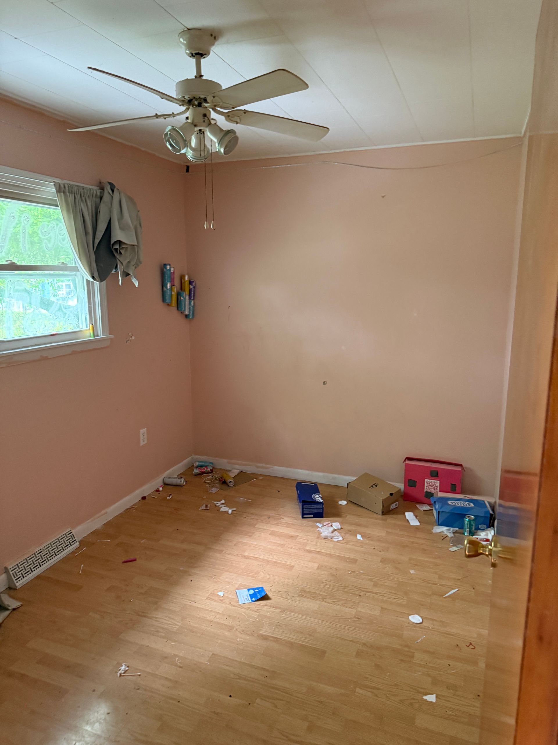 Empty room with peach walls, wood floor, ceiling fan, window with curtain.
