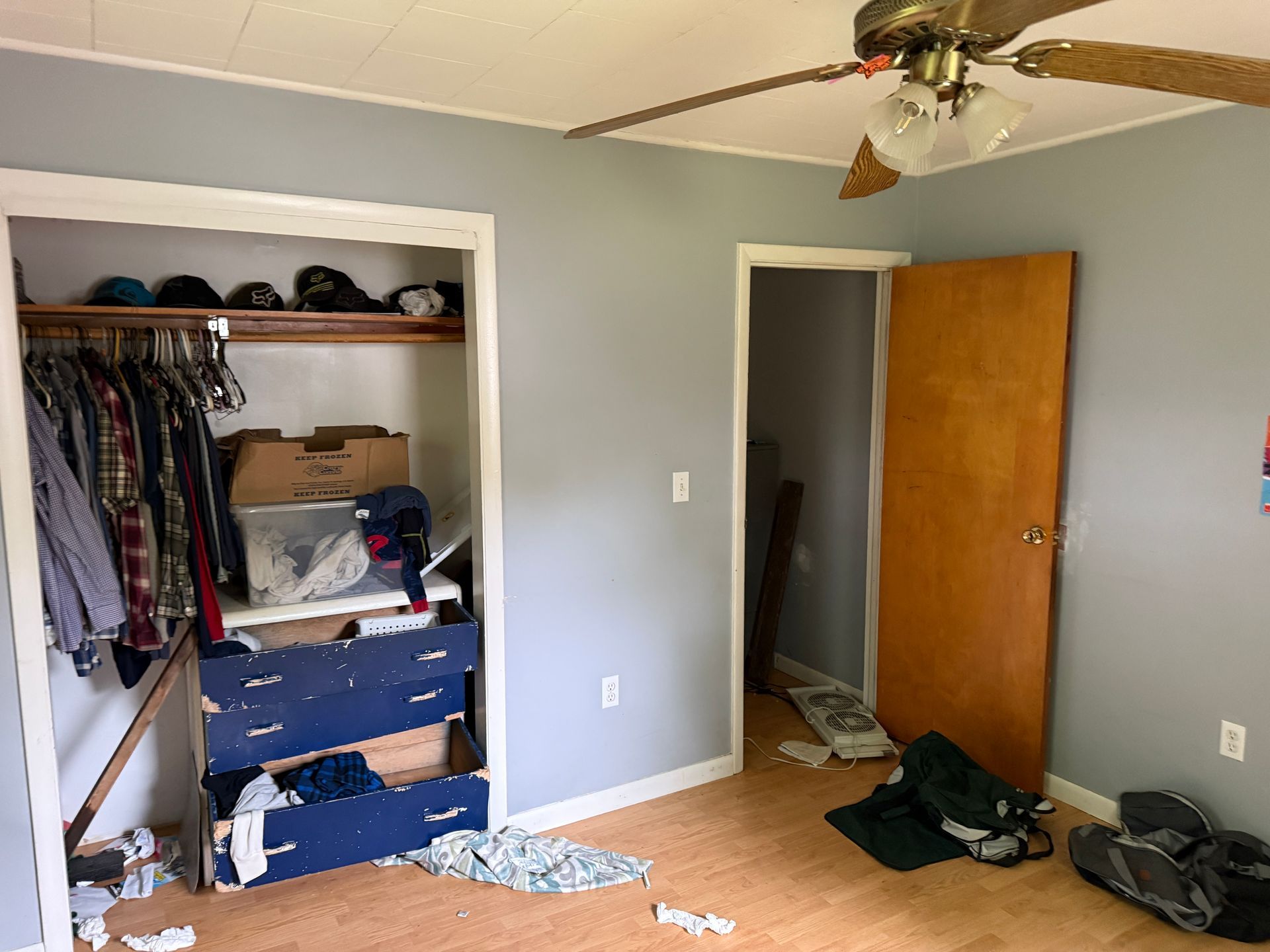 Bedroom interior with open closet, blue dresser, and wooden door. Grey walls, tan ceiling fan, and wood-look flooring.