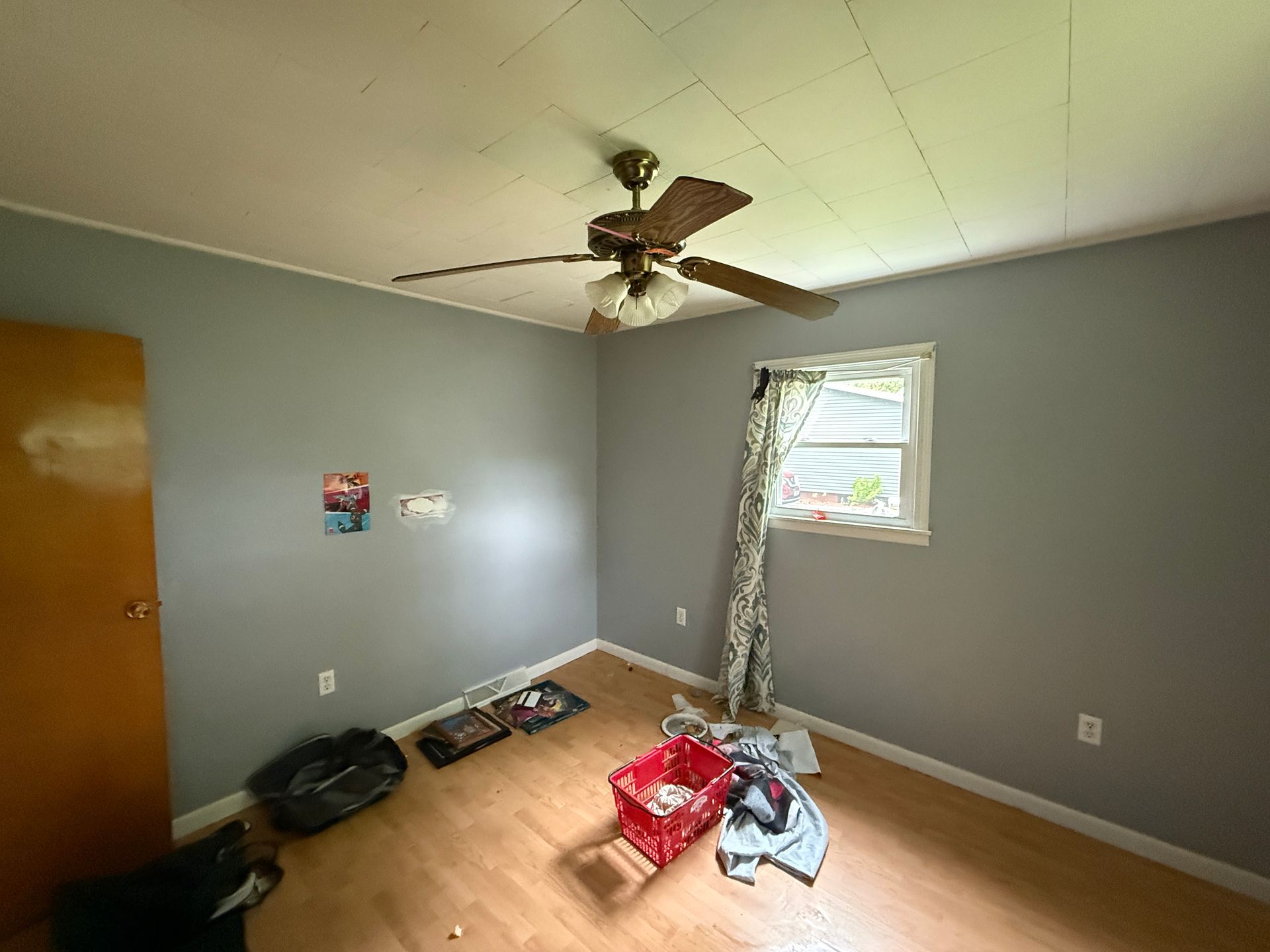 Empty room with gray walls, wooden floor, ceiling fan, and window with a curtain.