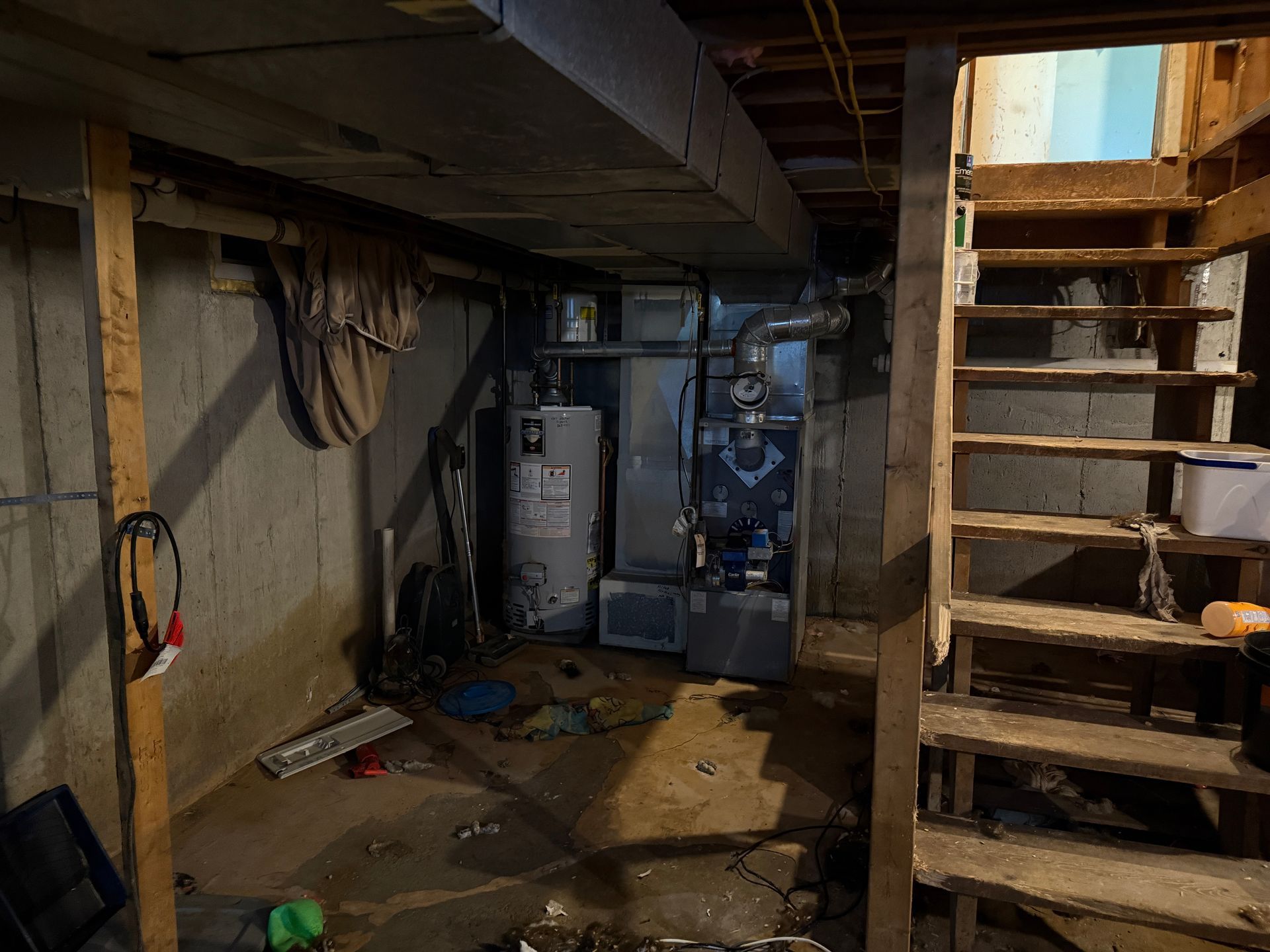 Basement with water heater, furnace, and wooden stairs; concrete walls, low ceiling.