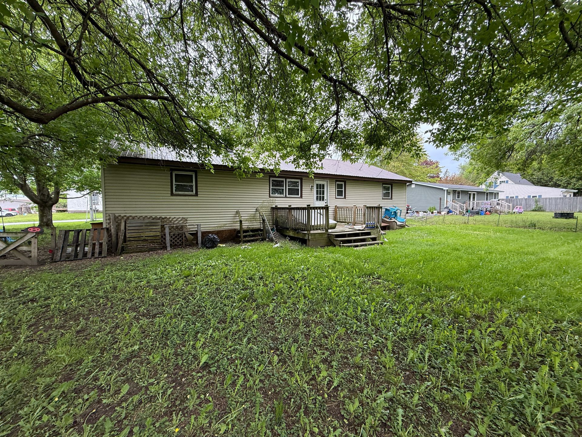 Backyard with grassy lawn, wooden deck, and tan house with several windows, beneath a tree.