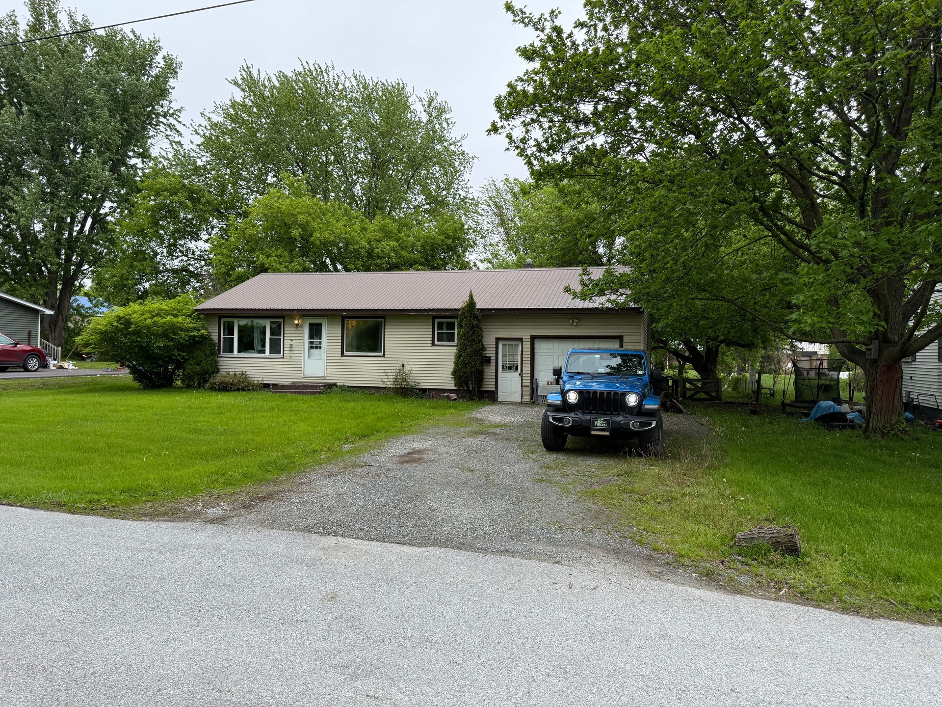 Tan house with attached garage, blue jeep parked in front. Gray gravel driveway. Green lawn, trees in background.