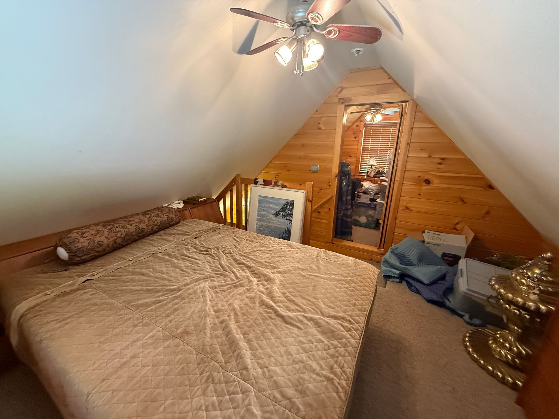 Bedroom in a cabin-style loft; bed with tan comforter, wooden walls, small closet, and a ceiling fan.