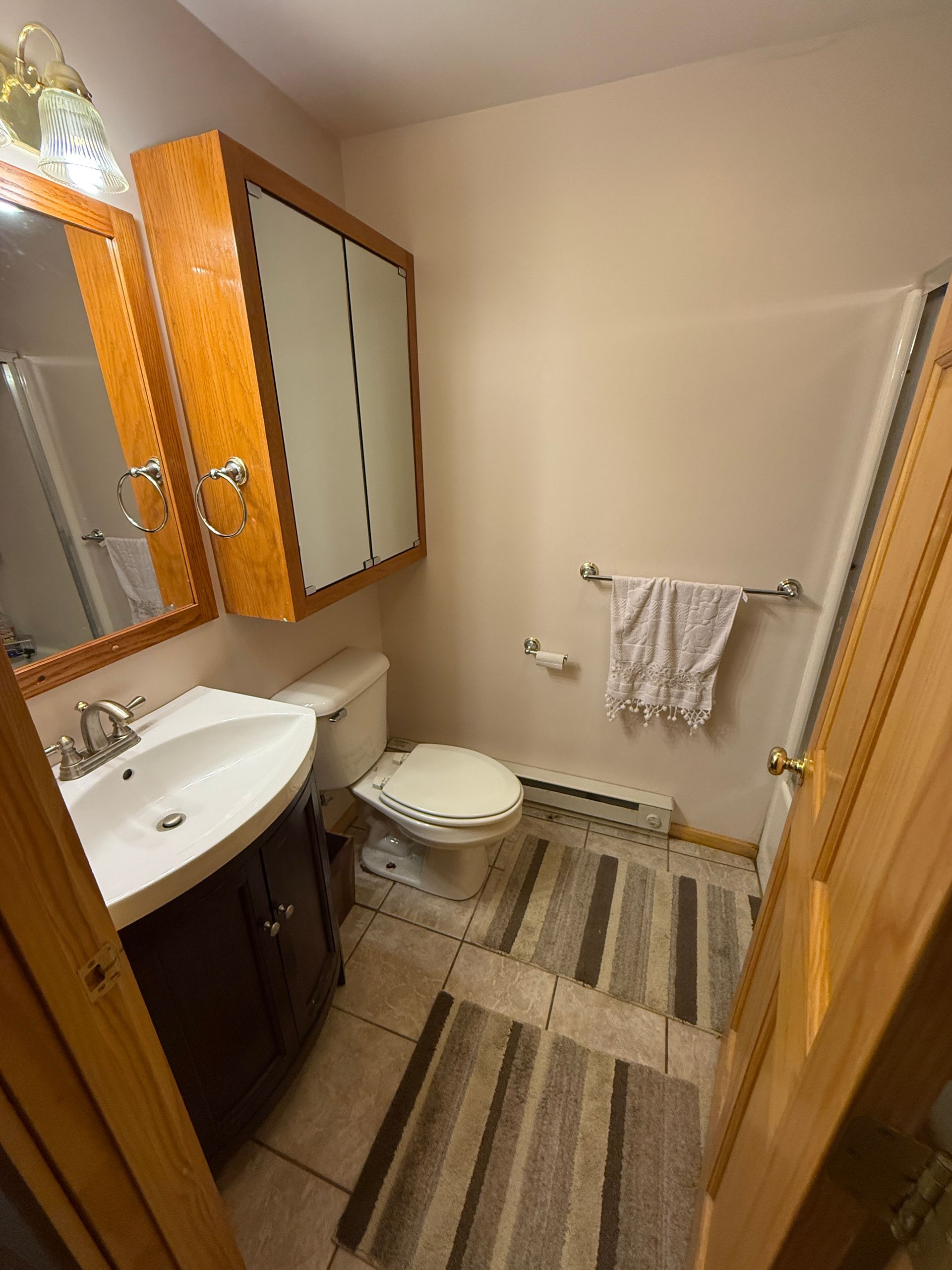 Small bathroom with a toilet, sink, and cabinets. Tan walls and brown floor mats.