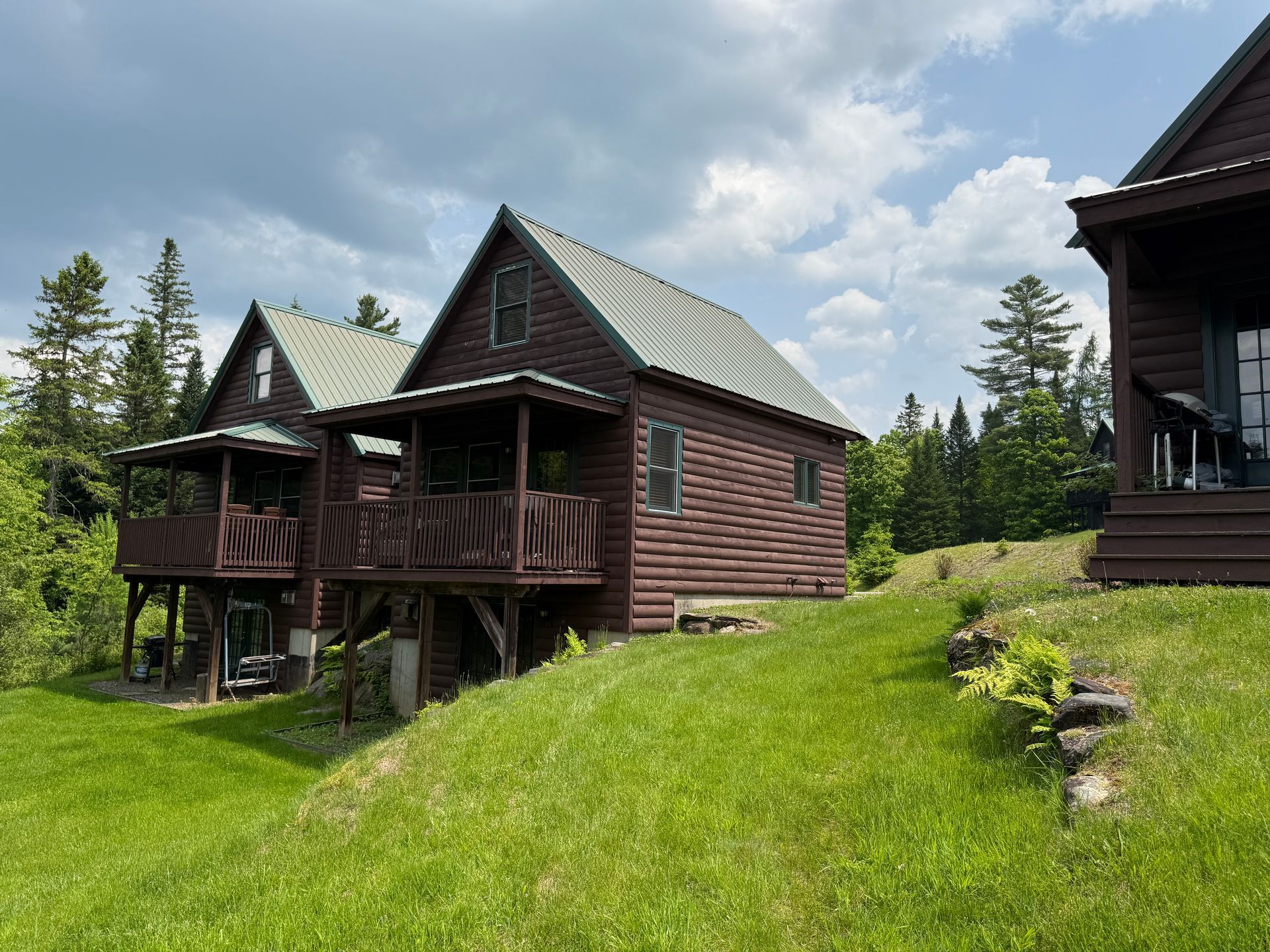 Wooden cabins with green metal roofs on a grassy hill under a cloudy sky.