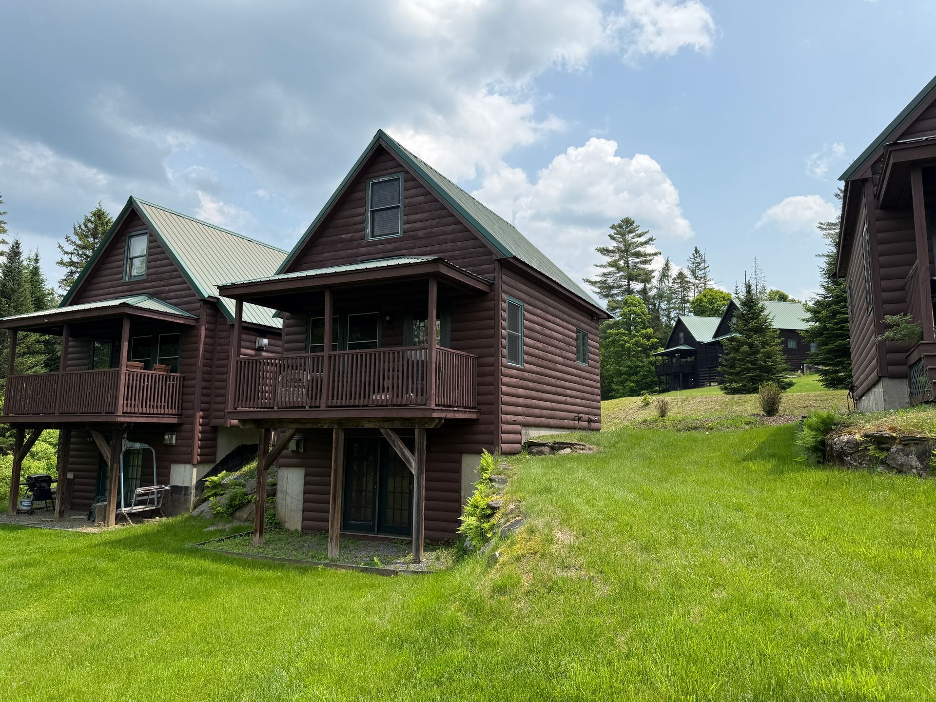 Wooden cabins with green roofs and brown porches on a grassy hillside under a cloudy sky.