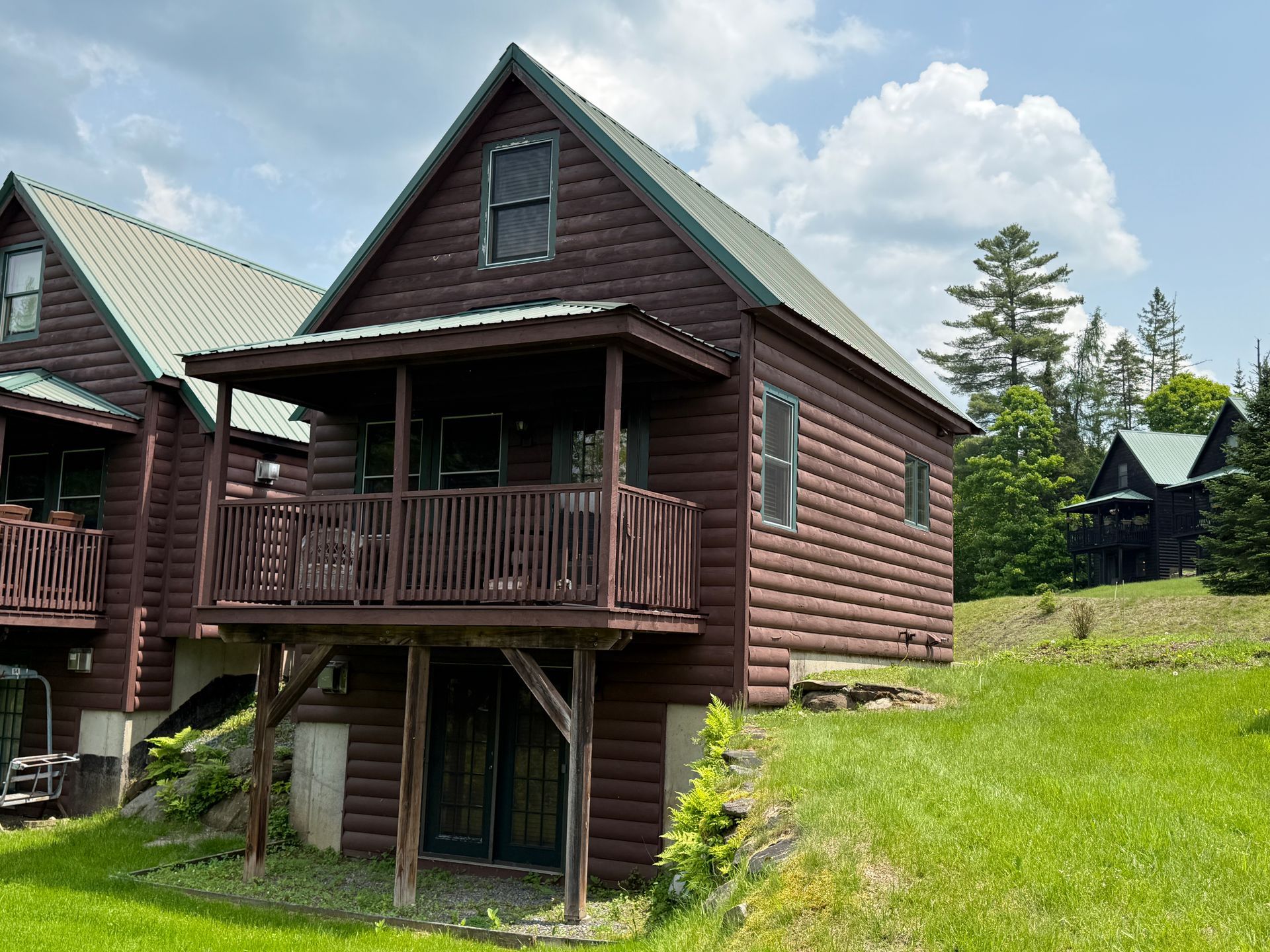 Brown cabin with a porch on a grassy hill under a partly cloudy sky.
