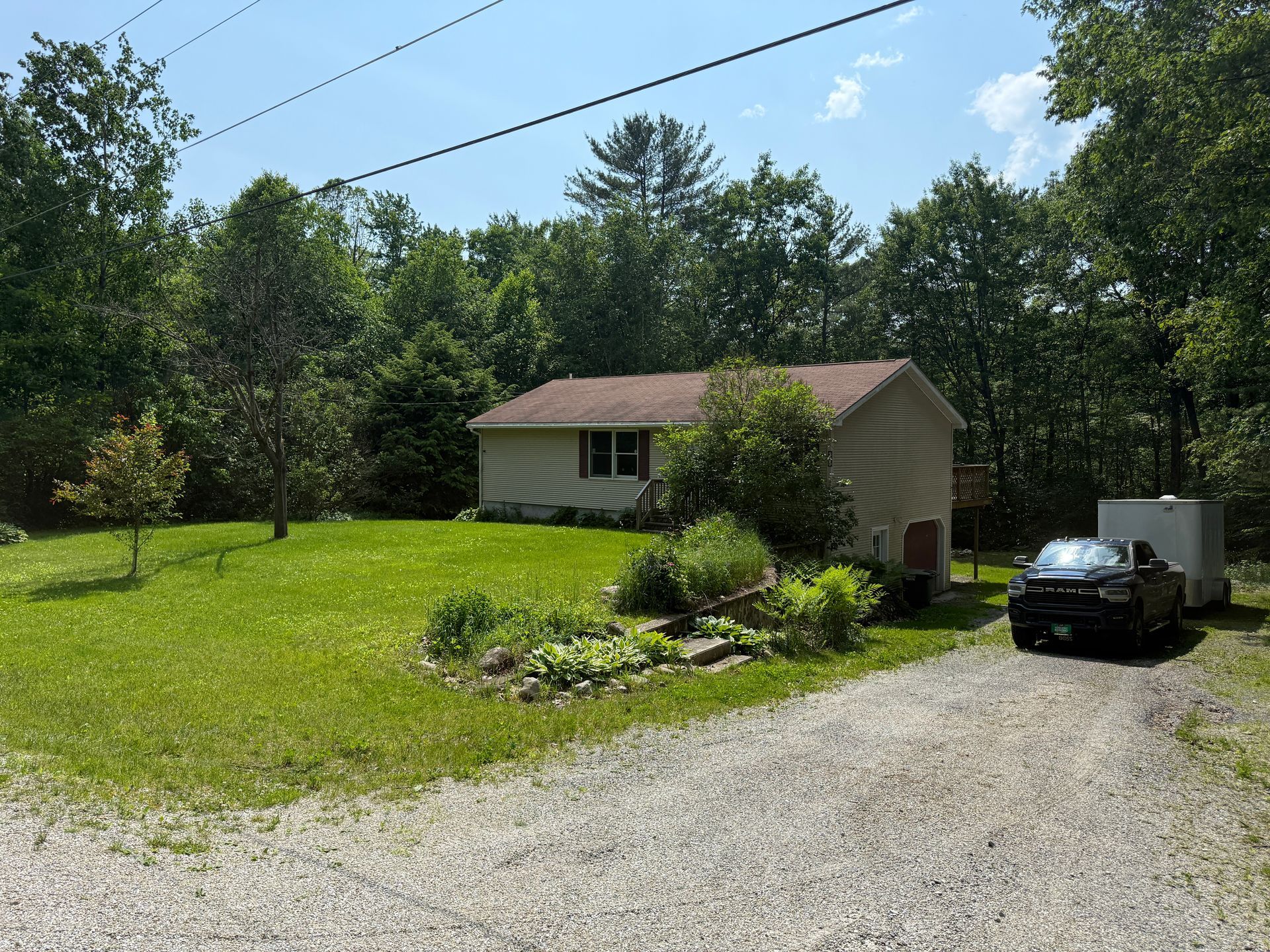 Small beige house with brown roof and gravel driveway in a wooded area; car parked next to trailer.