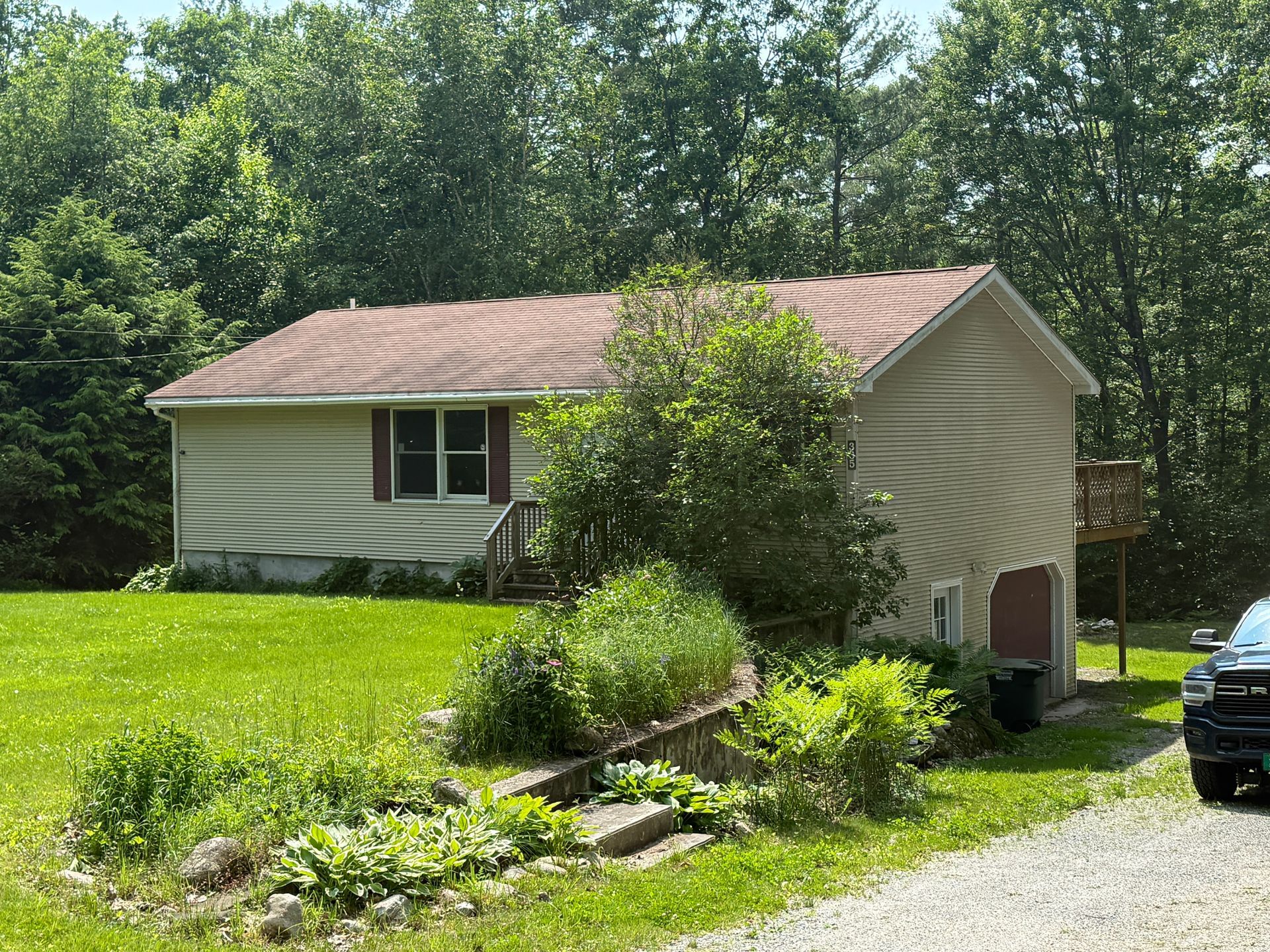 Small beige house with brown roof and green lawn, surrounded by trees.