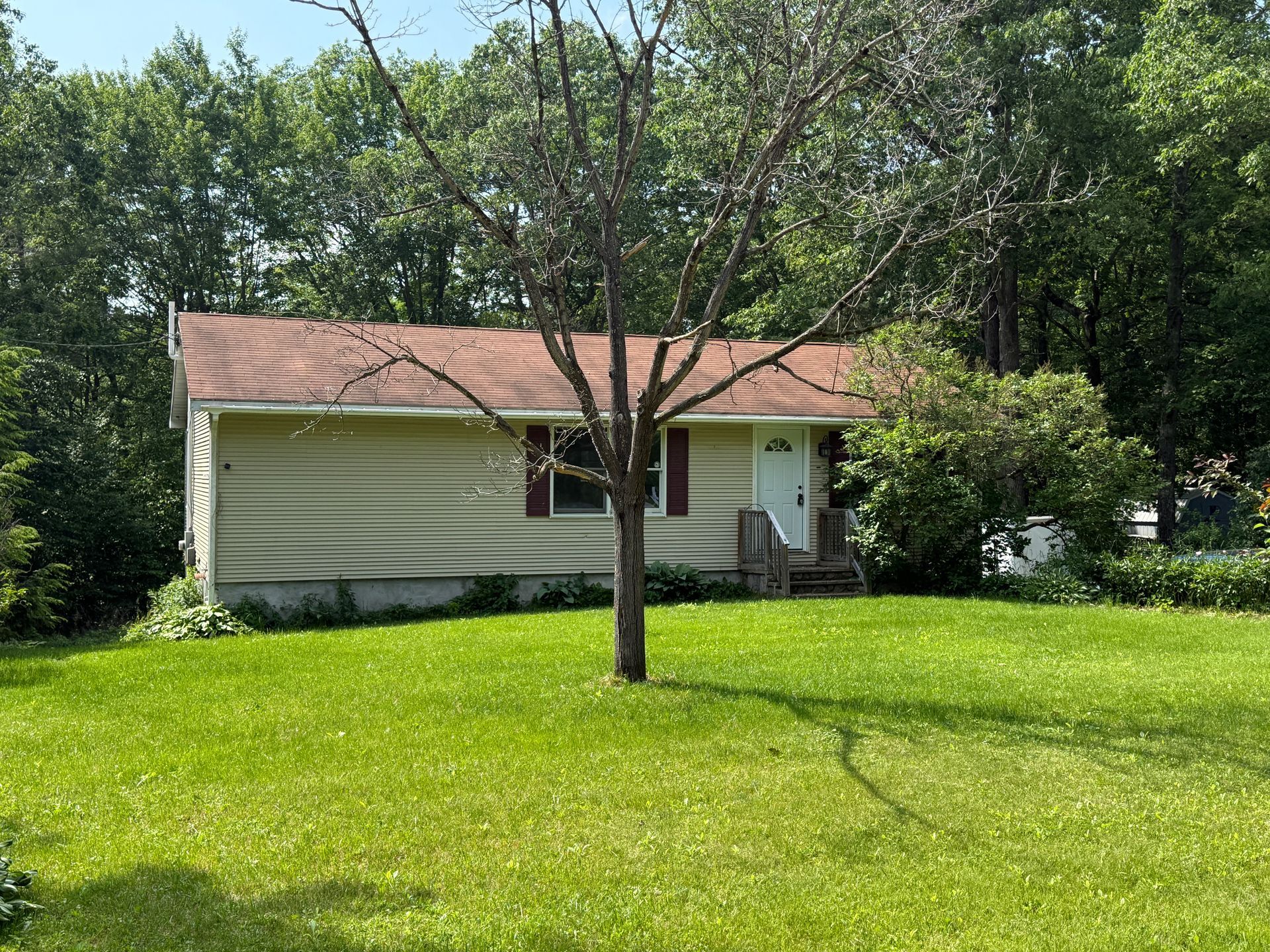 Small yellow house with a brown roof and red shutters, surrounded by green grass and trees.