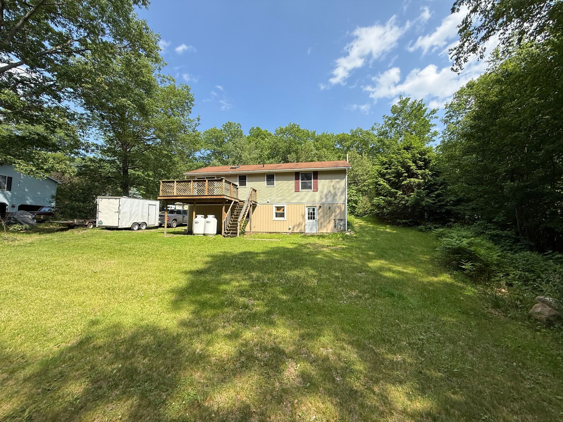 Two-story house with wooden deck and staircase, on a grassy lot surrounded by trees under a blue sky.