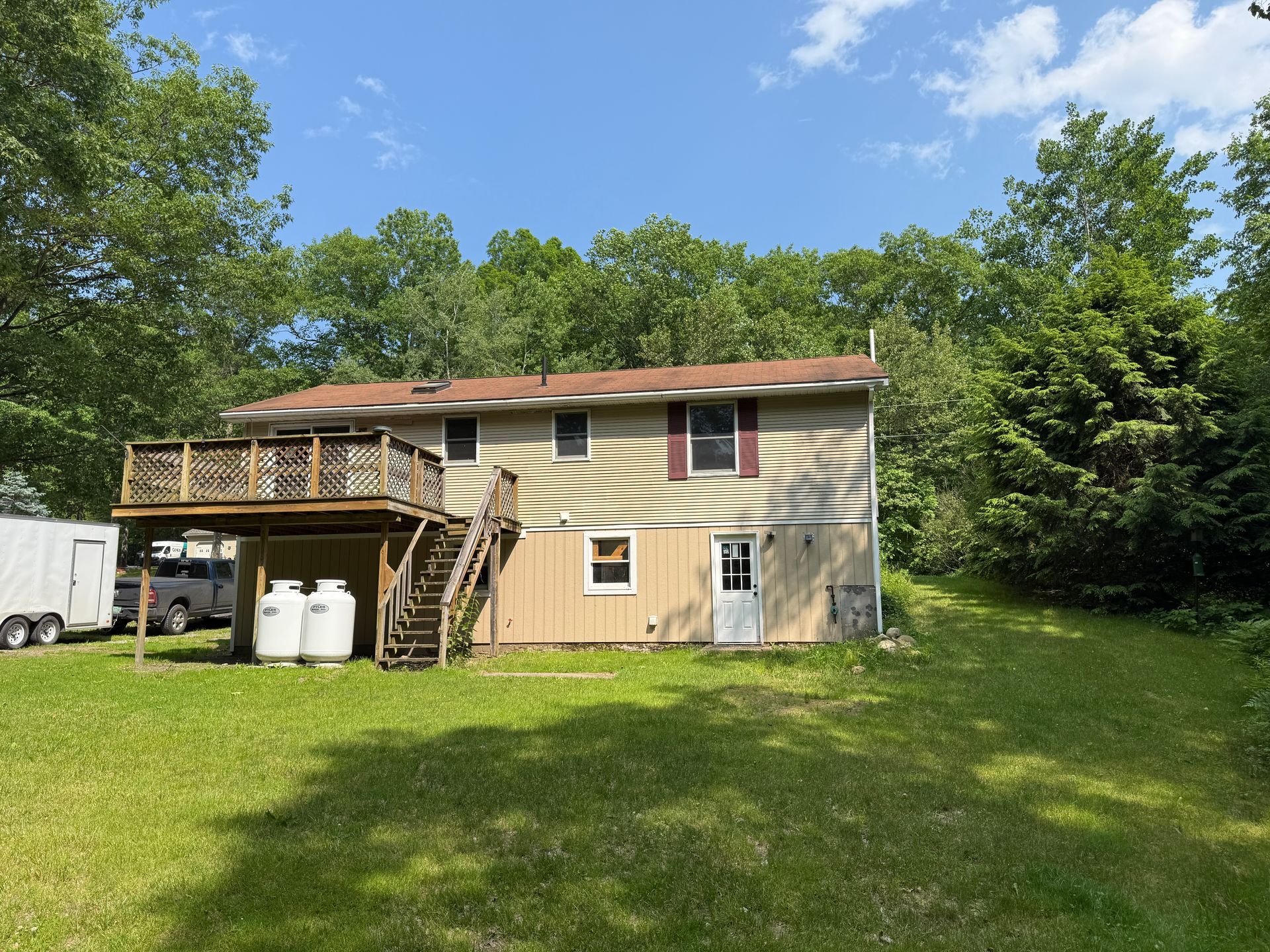 Back of a two-story beige house with deck and stairs. Two propane tanks sit below the deck on the grassy lawn.
