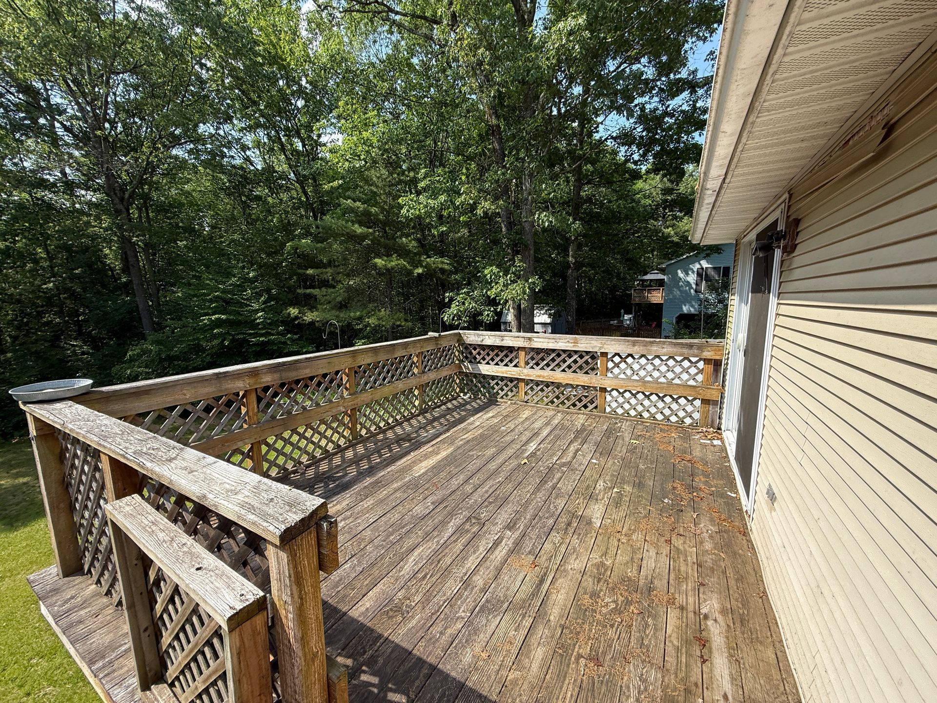Wooden deck attached to a house with latticework railings. Trees in the background, sunny day.