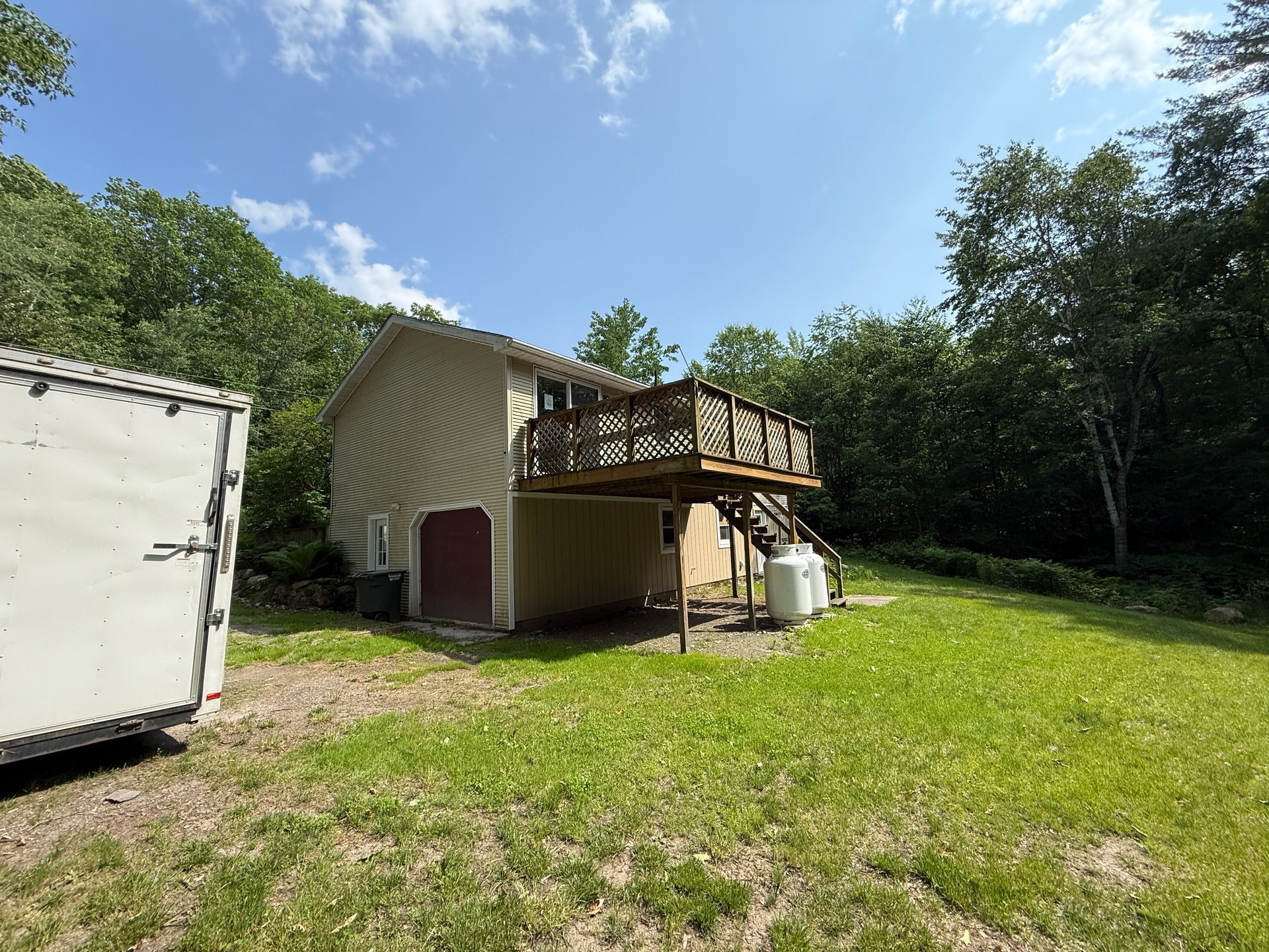 Two-story house with a wooden deck, propane tanks, and a trailer on a grassy lot surrounded by trees.