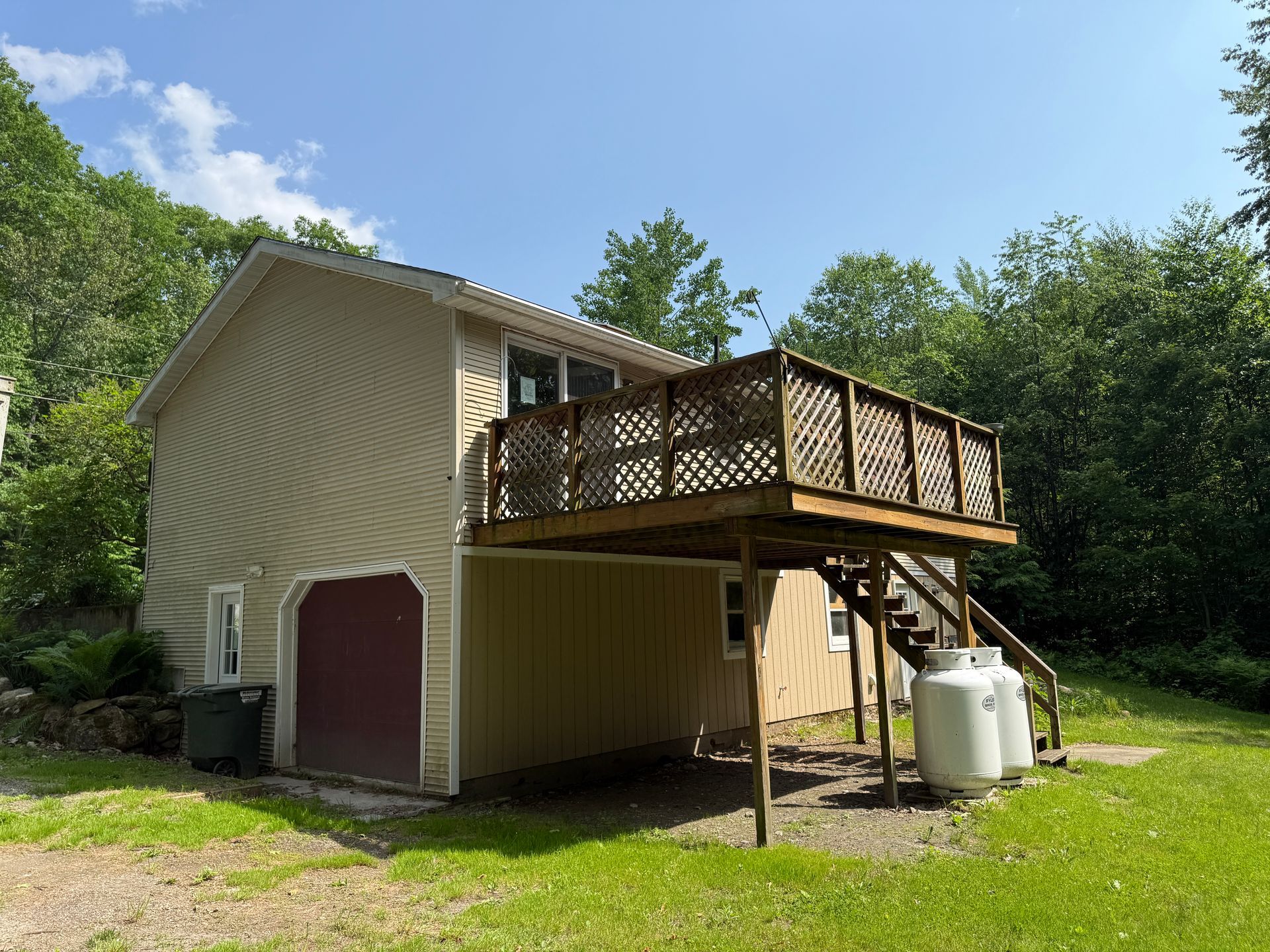 Two-story building with a deck, propane tanks, and a garage door, set in a grassy area with trees and a blue sky.