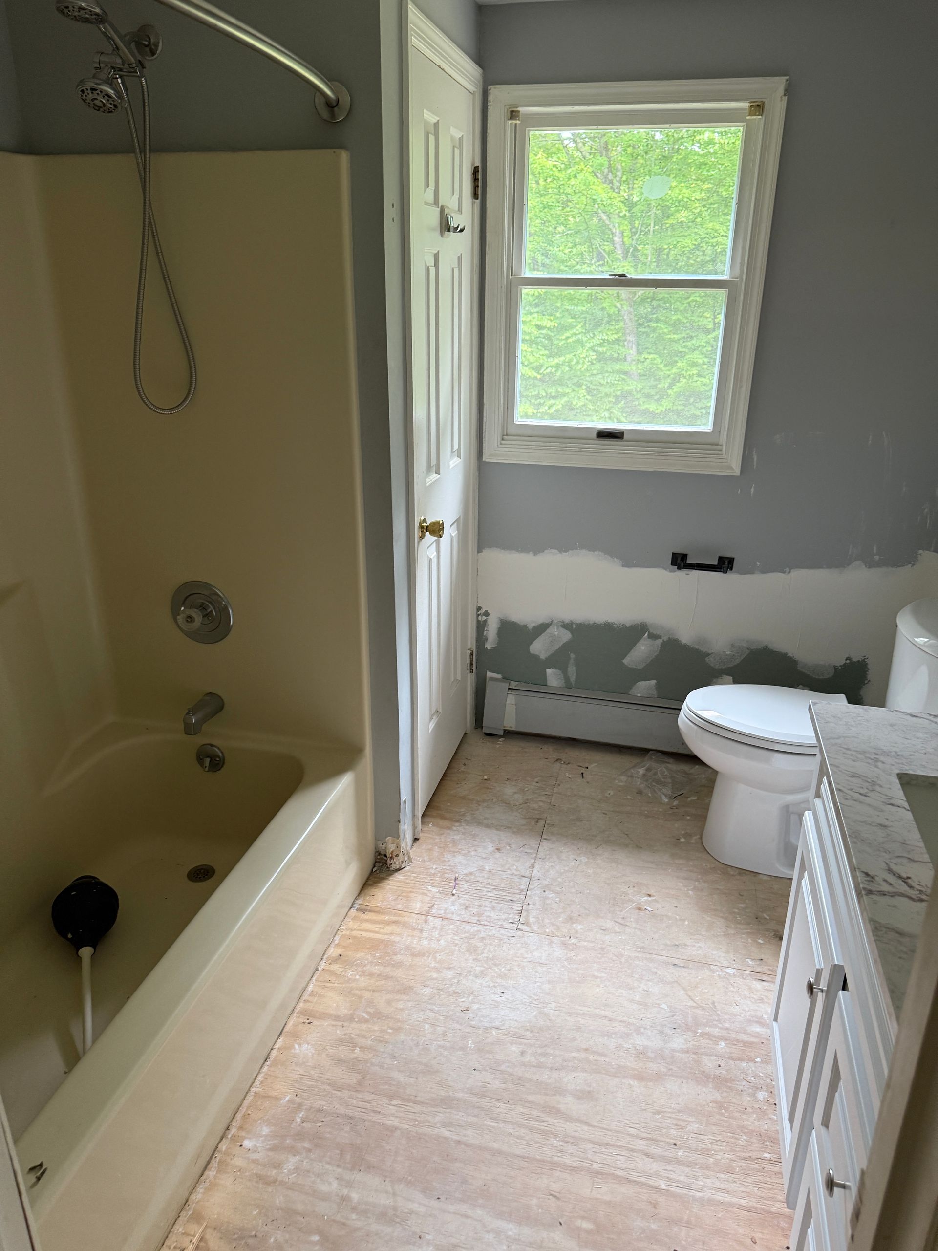Bathroom with off-white bathtub, toilet, and vanity; window with greenery outside; blue-gray walls, and beige flooring.