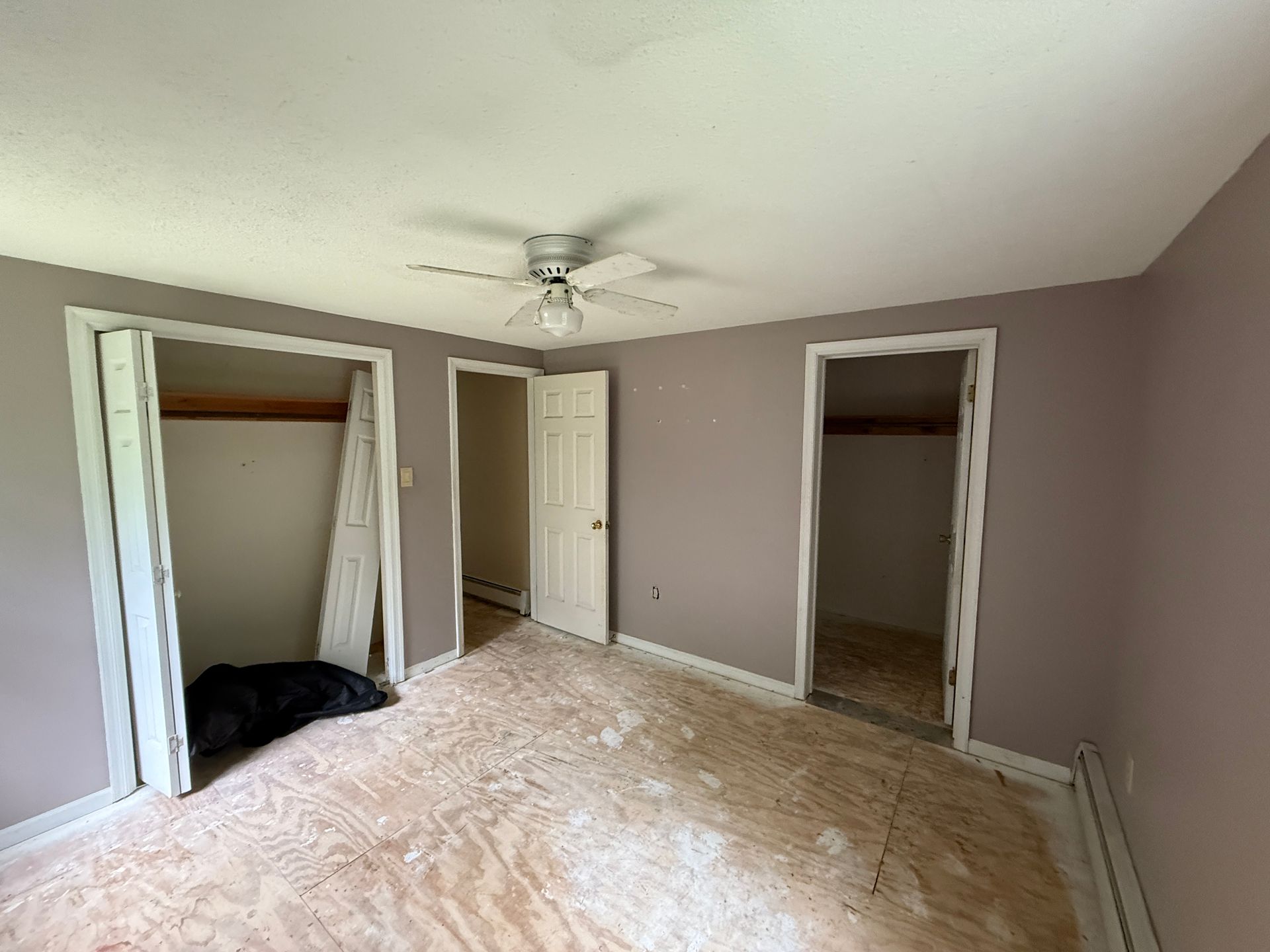 Empty bedroom with plywood floor, three doorways, purple walls, and ceiling fan.