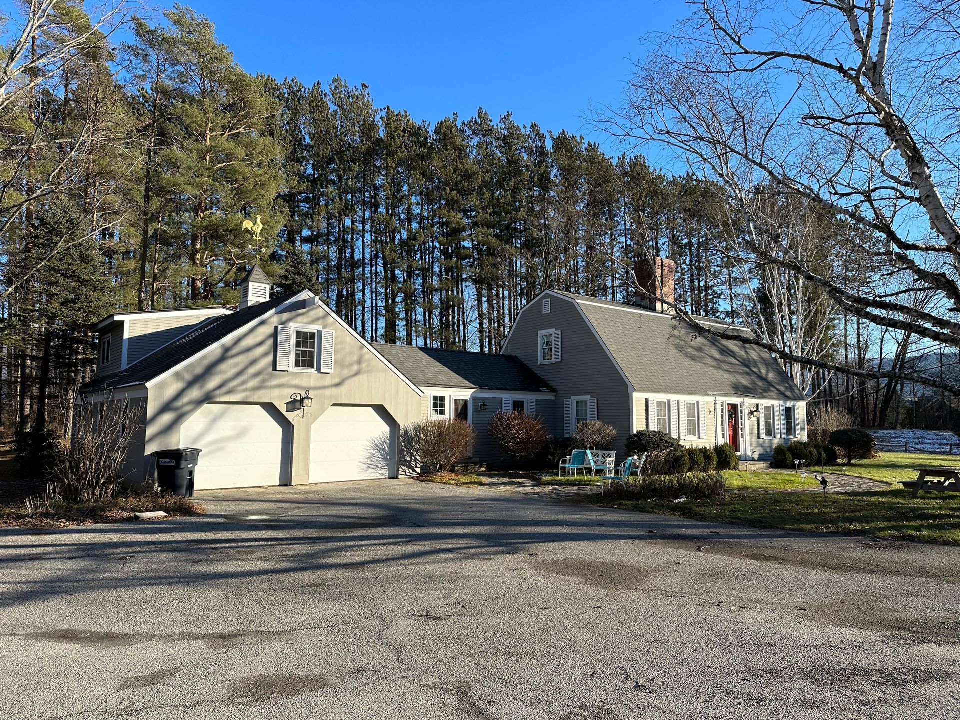 Gray house with attached garage, tall trees in the background, and a gravel driveway.