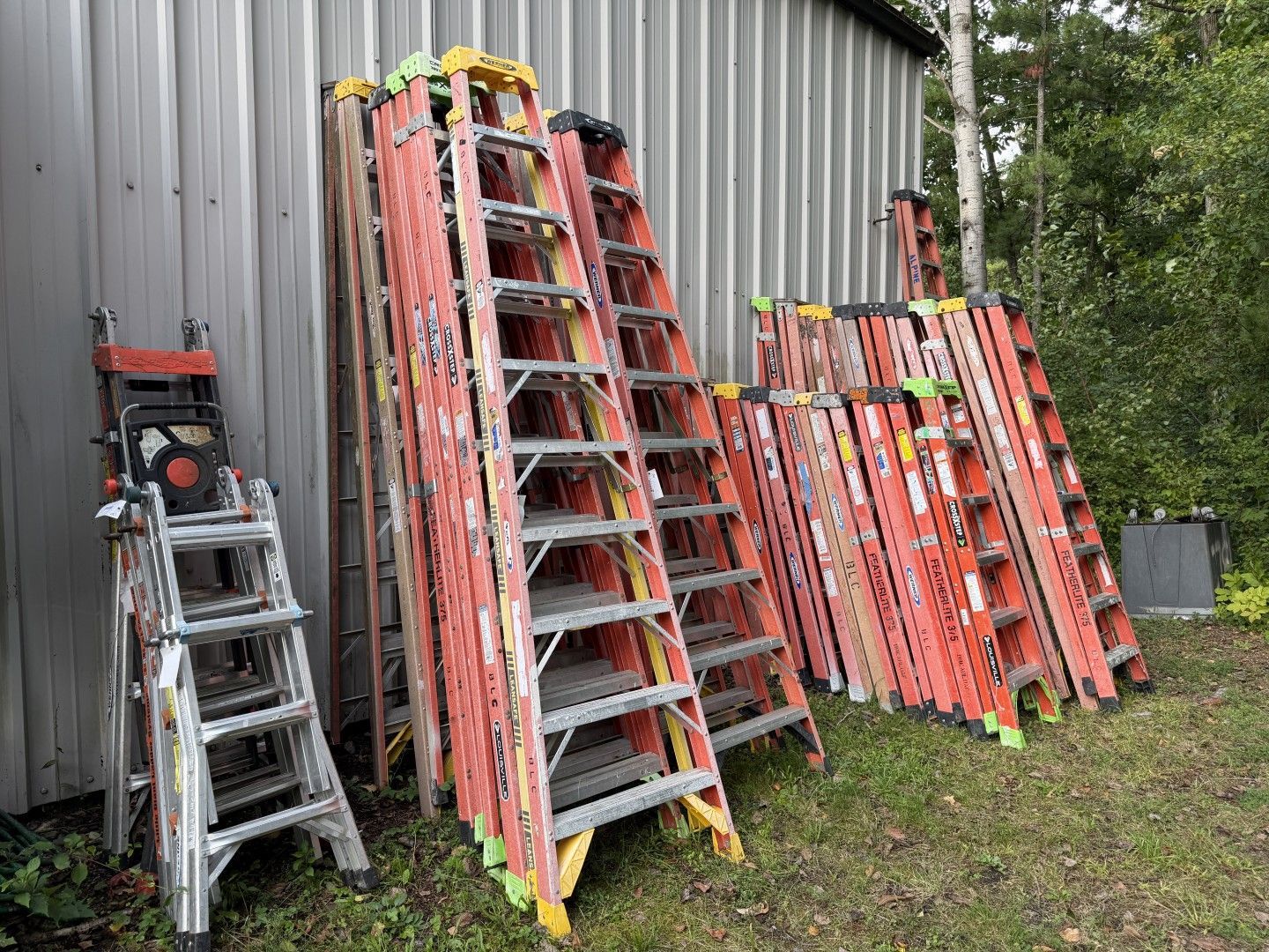 Stacks of orange and silver extension ladders leaning against a corrugated metal building outdoors