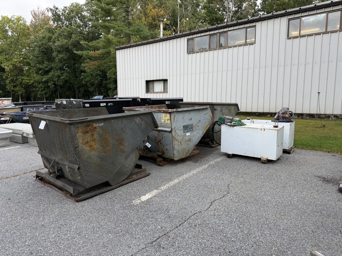 Large metal dumpsters and a white storage bin on gravel beside a white industrial building
