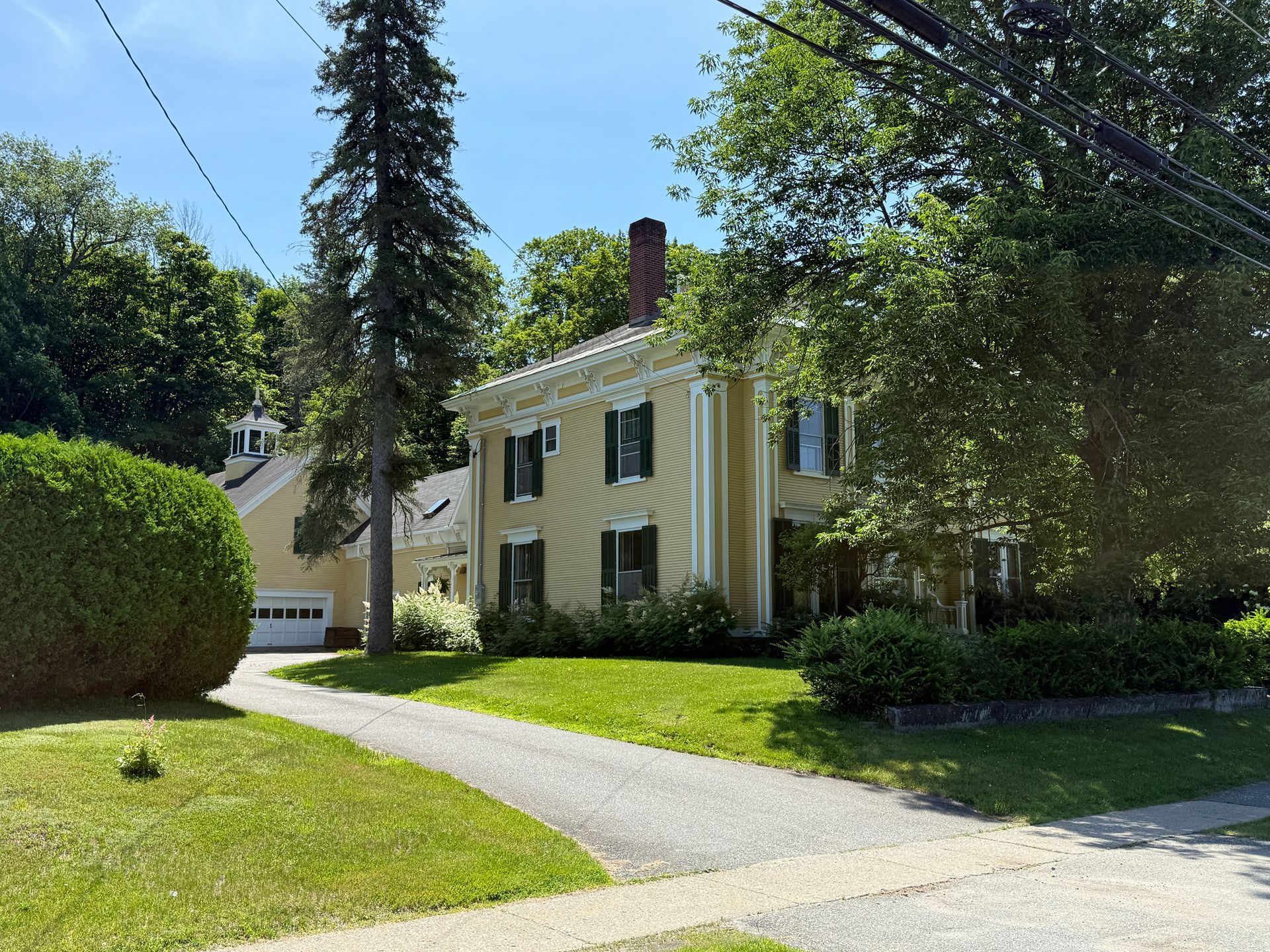 Yellow historic house with columns, green shutters, and a driveway, on a sunny day.