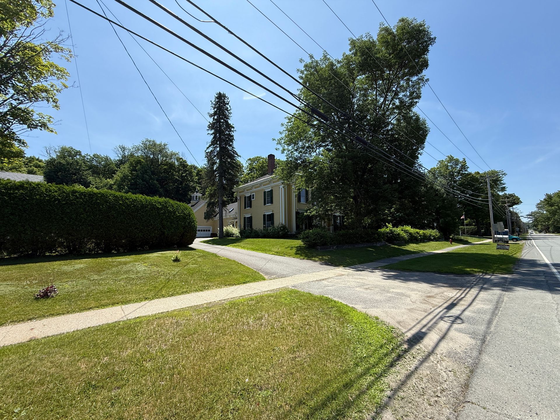 Residential street with houses, trees, power lines, and green lawns on a sunny day.