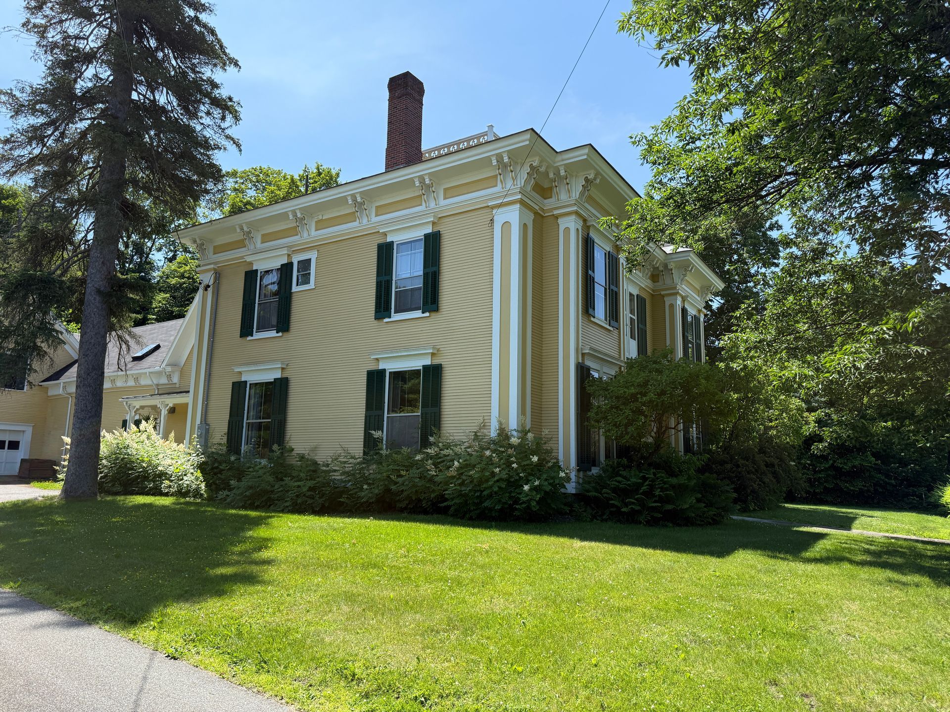 Yellow house with white trim, green shutters, and a brick chimney on a sunny day.