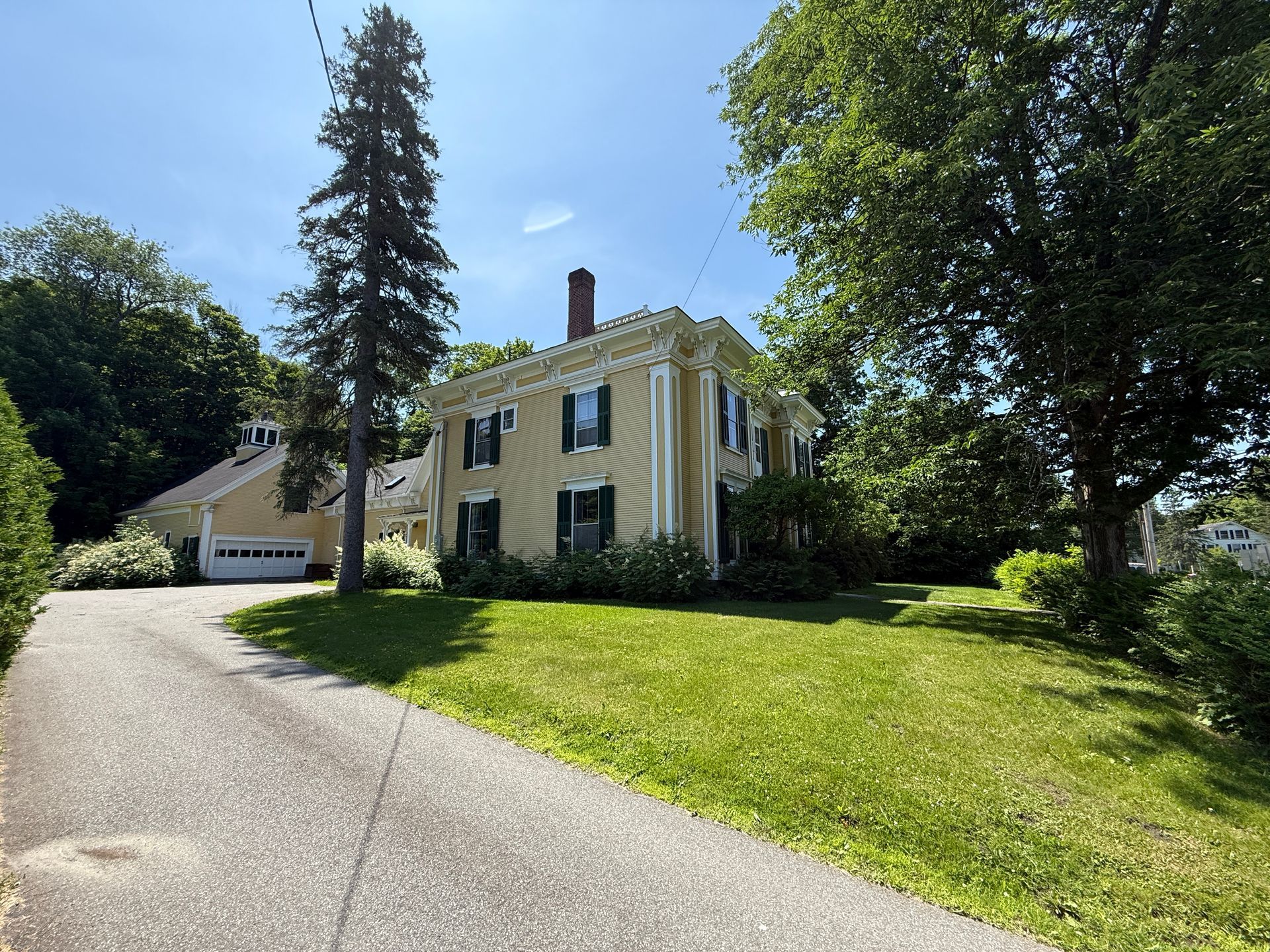 Yellow house with columns, green lawn, driveway. Blue sky, trees surround.