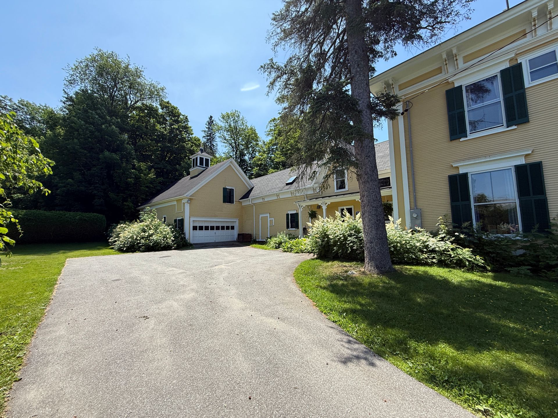 Yellow house with a driveway and garage, surrounded by trees and greenery.