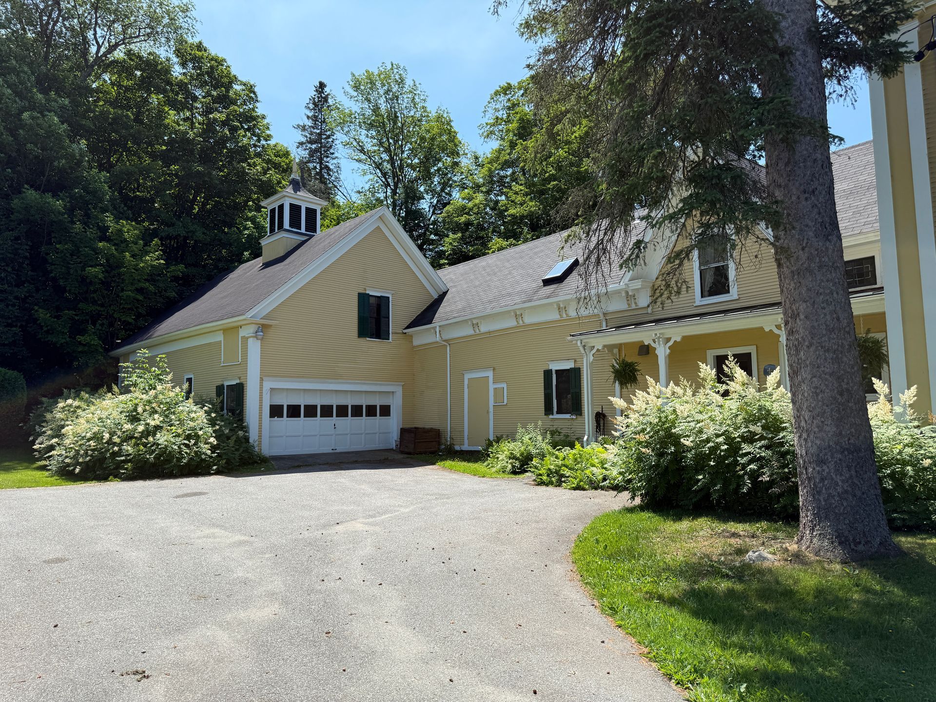 Yellow house with a garage and a gravel driveway. Green bushes and trees surround the building. Blue sky.
