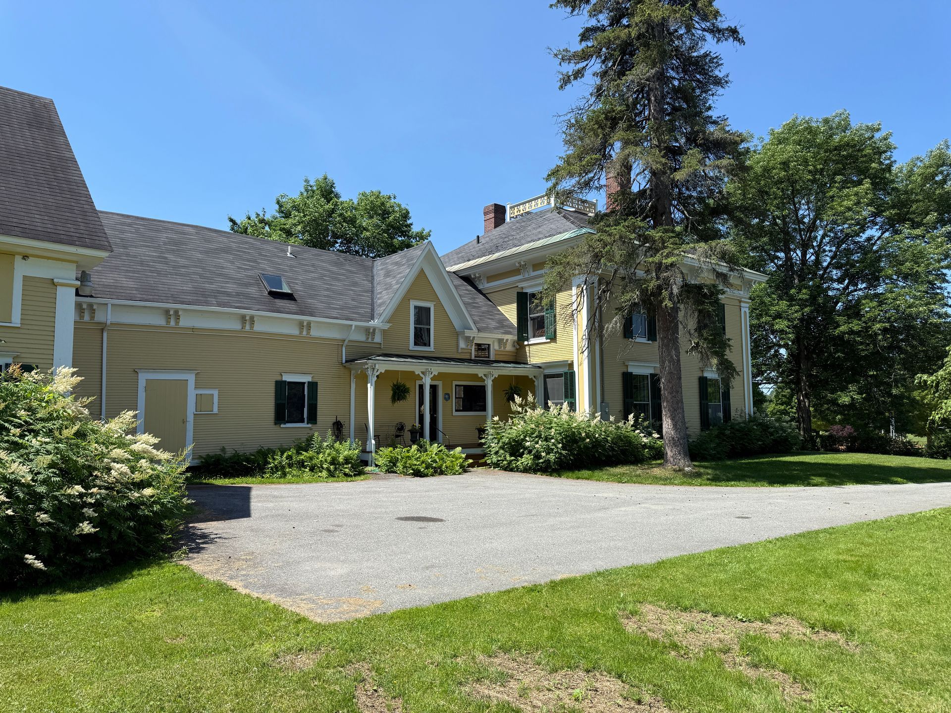 Yellow two-story house with black shutters, porch, and a gravel driveway under a blue sky.