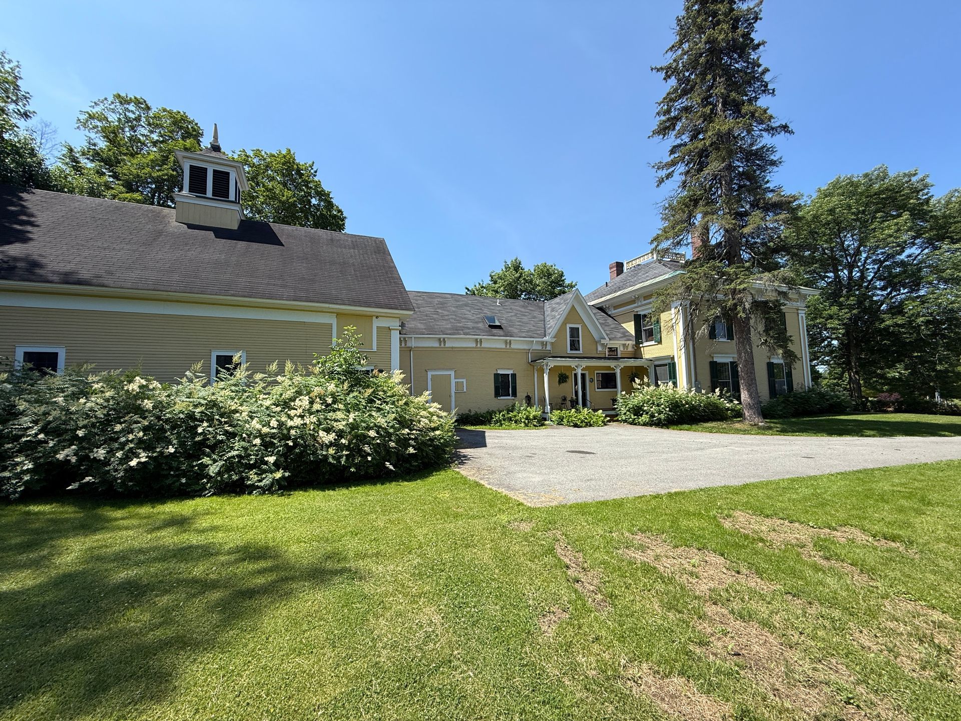 Yellow house and connected barn, surrounded by green grass and trees, under a blue sky.