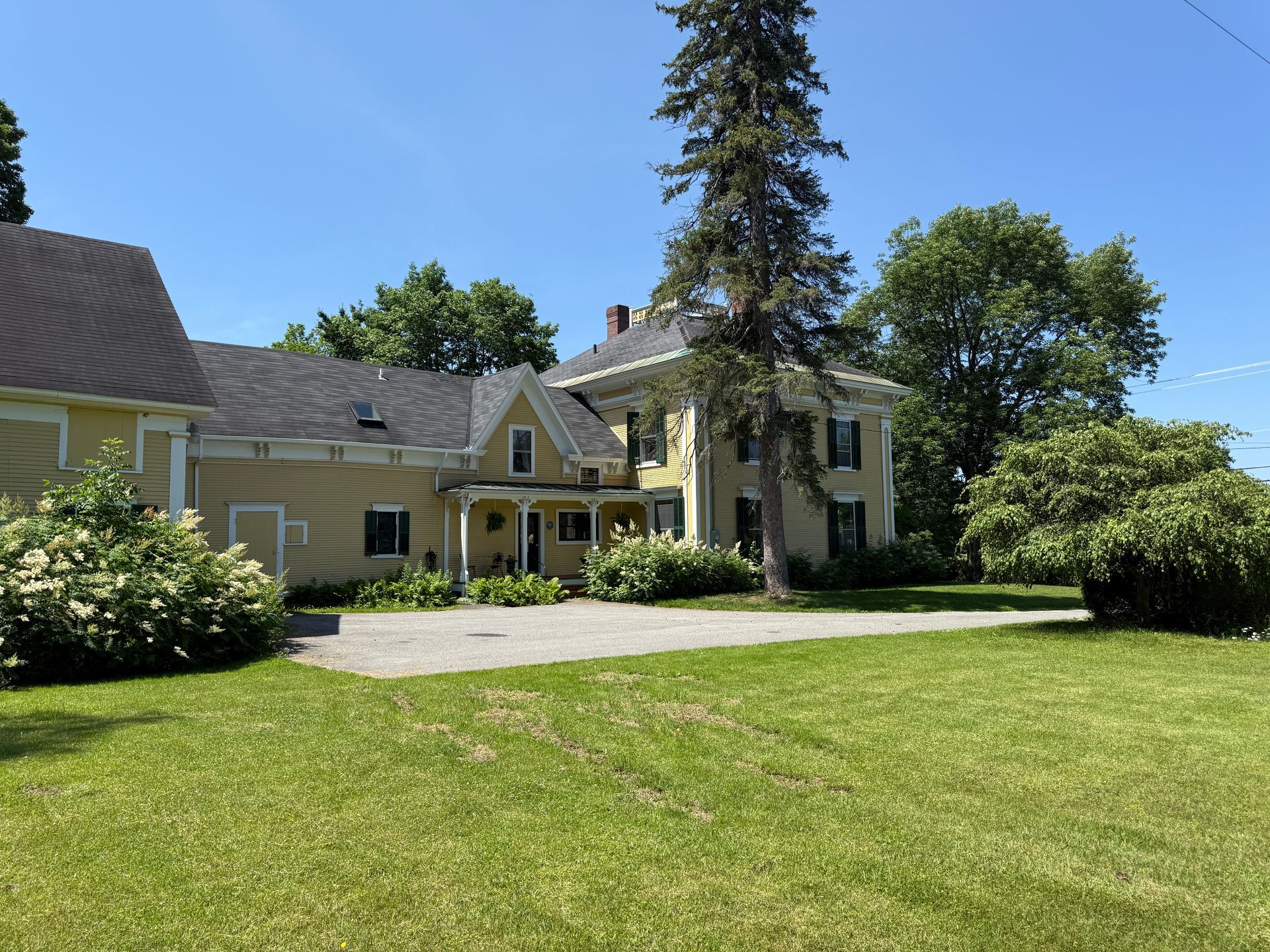 Yellow house with a gravel driveway, surrounded by green grass and trees under a blue sky.