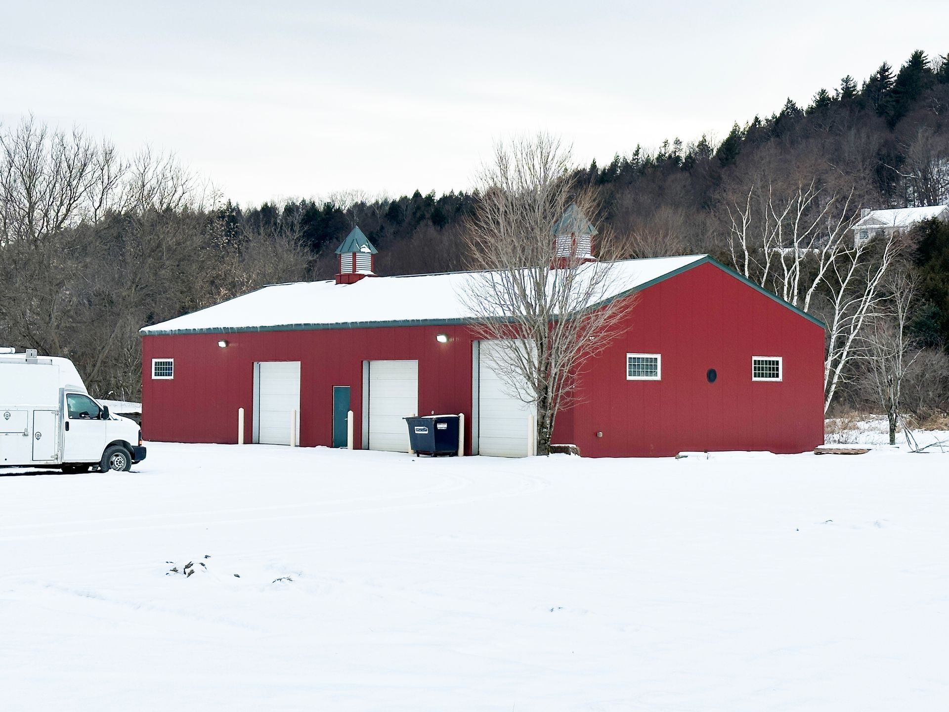 Red building with three garage doors in a snow-covered field, evergreen trees in the background.