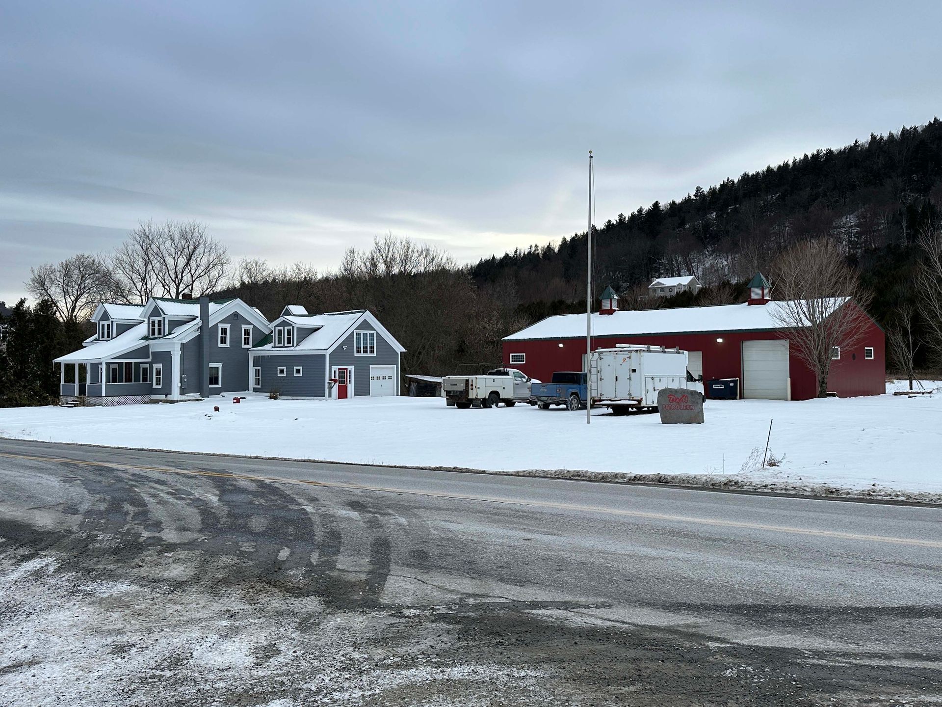 Snow-covered farm scene with gray house, red barn, and bare trees under a cloudy sky.