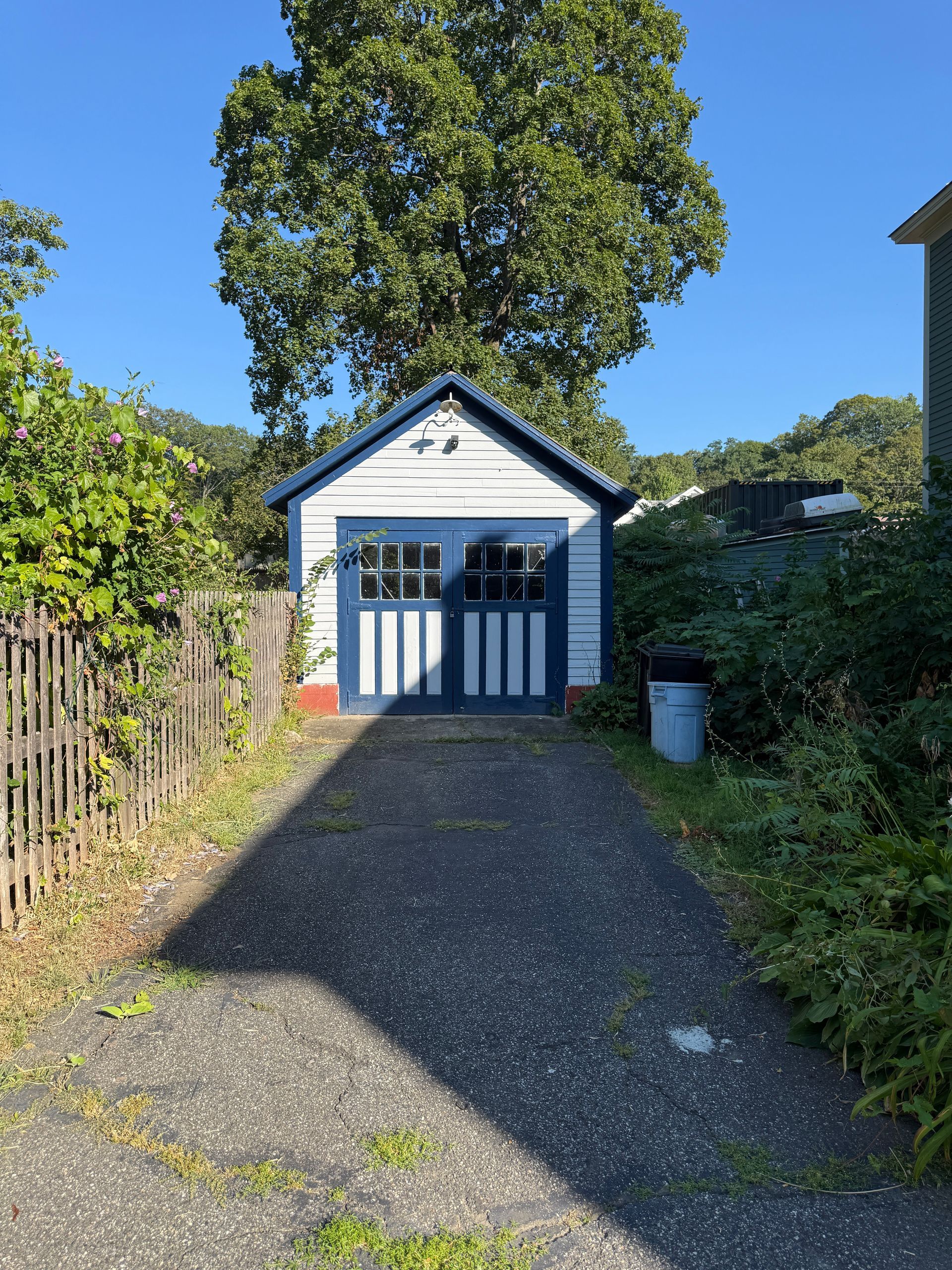 Small blue and white garage with a driveway, set against a backdrop of trees and greenery.