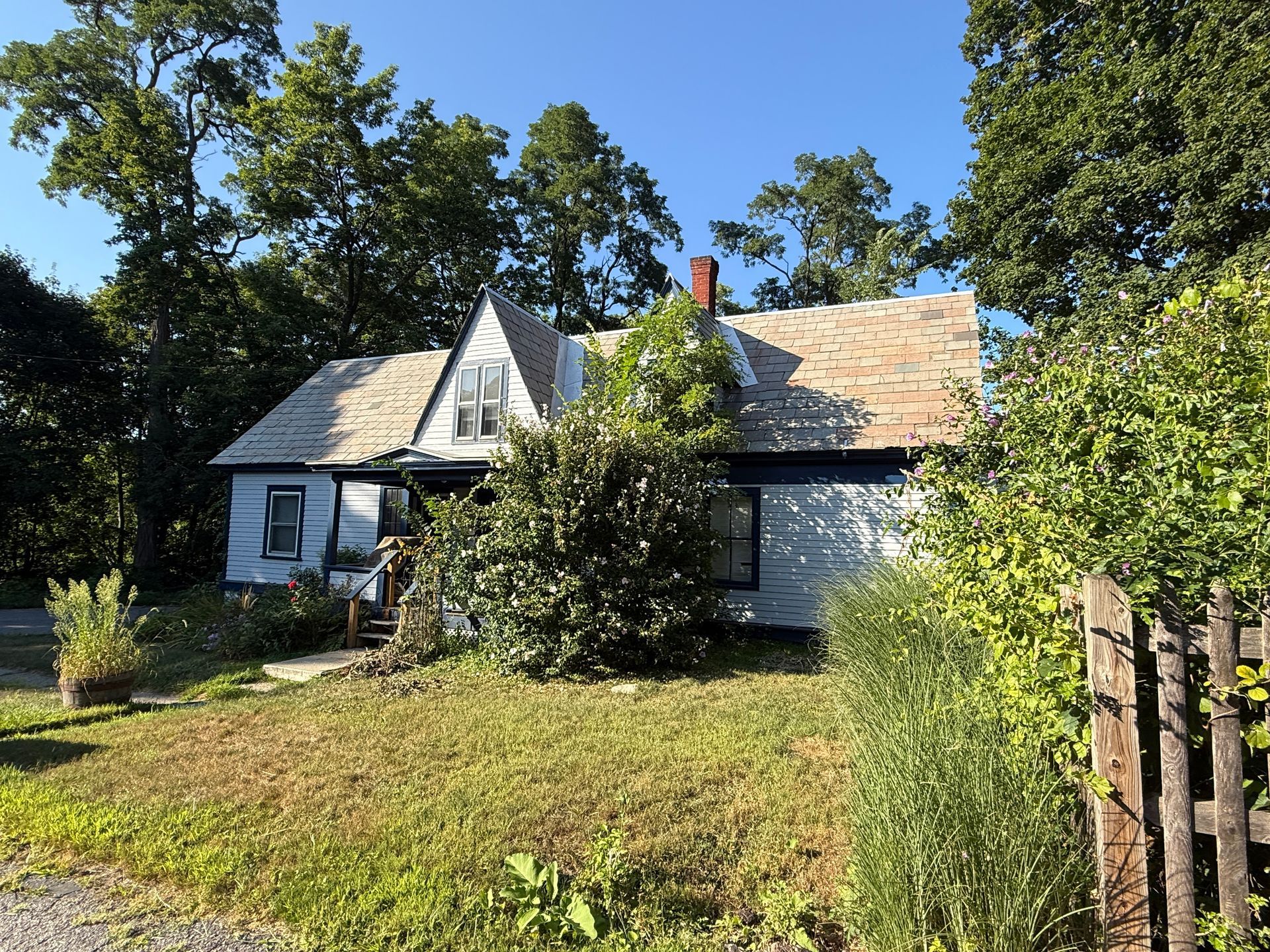 Small, light-colored house with a dark blue trim and a brown roof, surrounded by green foliage and trees.