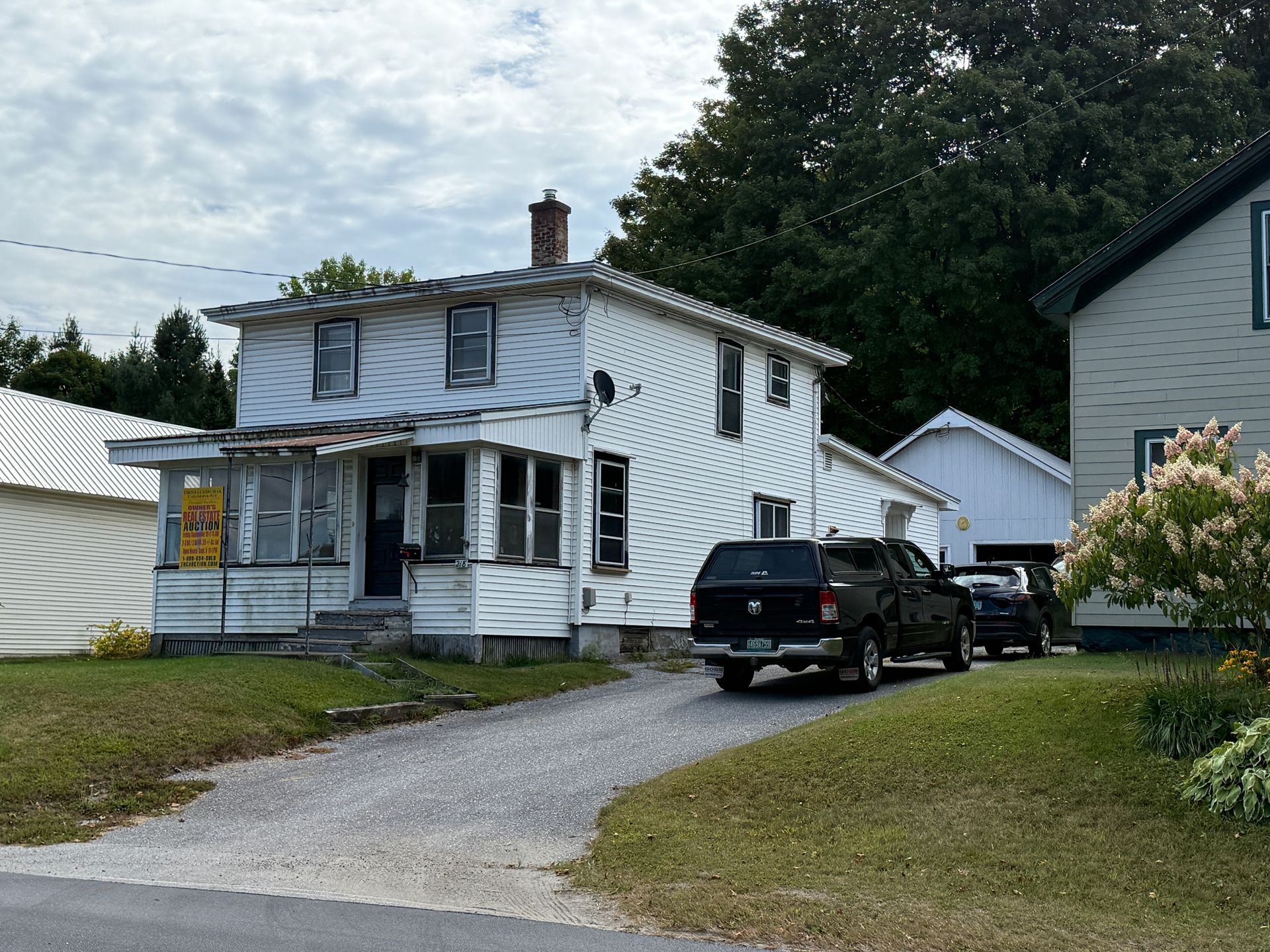 White two-story house with porch, driveway with vehicles, overcast sky, trees in the background.