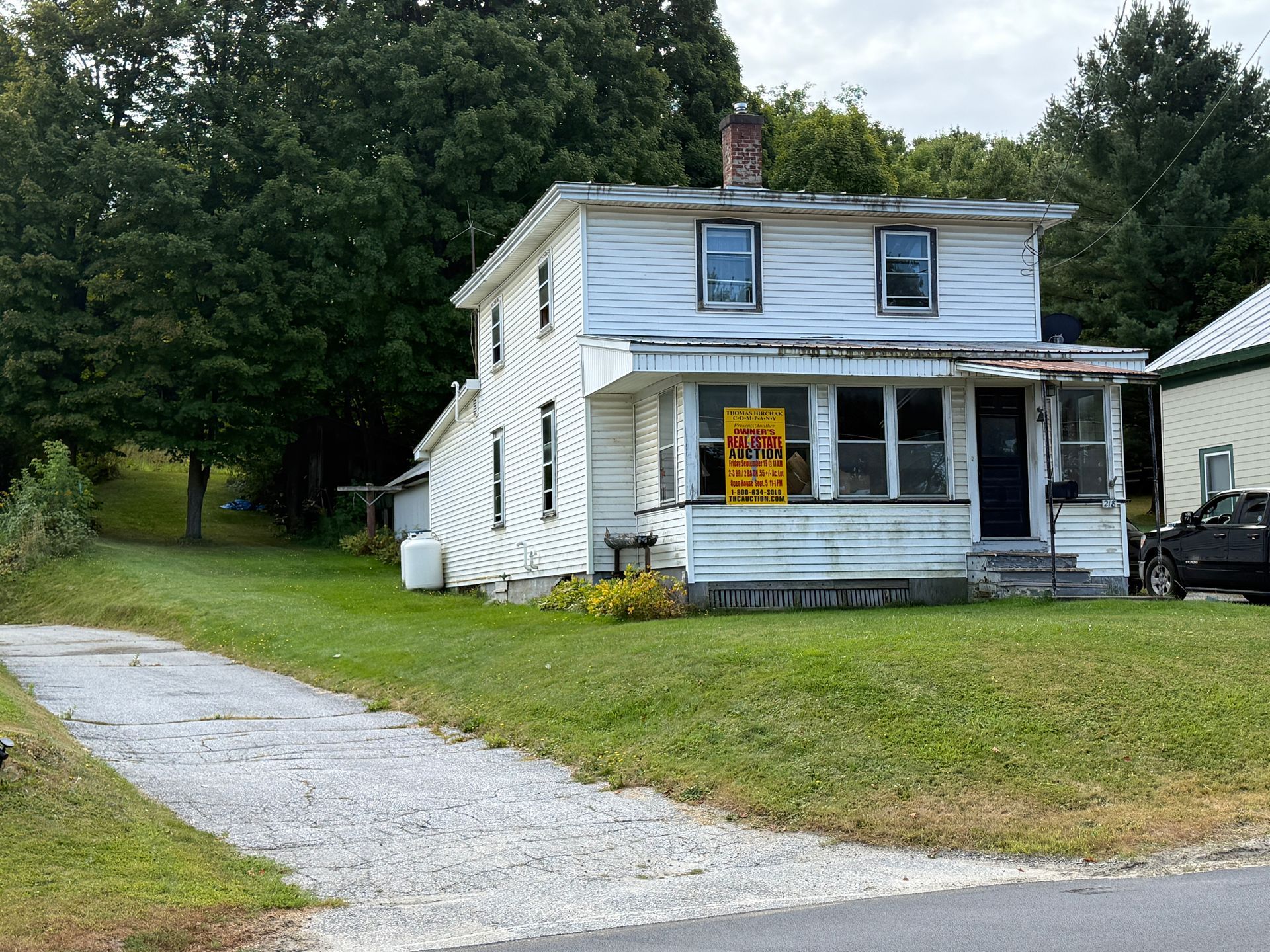 White two-story house with porch; gravel driveway; tall trees in background; a 