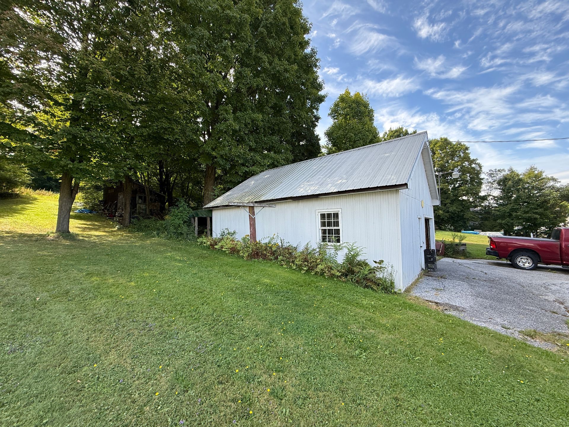 White barn with metal roof on grassy hill, trees in background, red truck parked nearby.