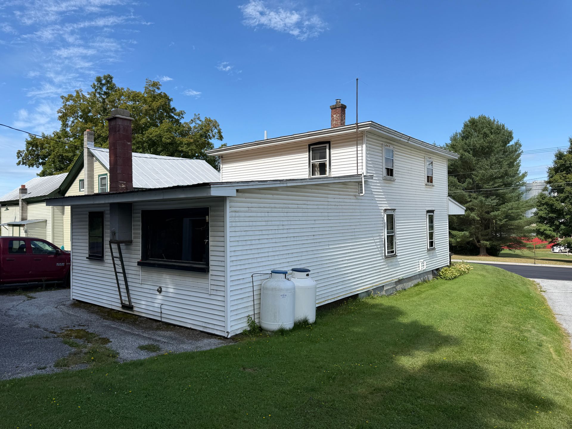White two-story house with attached single-story section, propane tanks, and chimney under a blue sky.