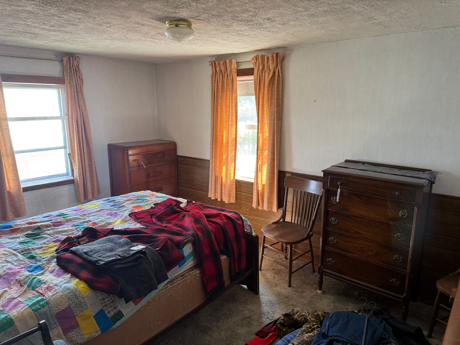 Bedroom with bed, dressers, chair, windows with curtains; walls are panelled.