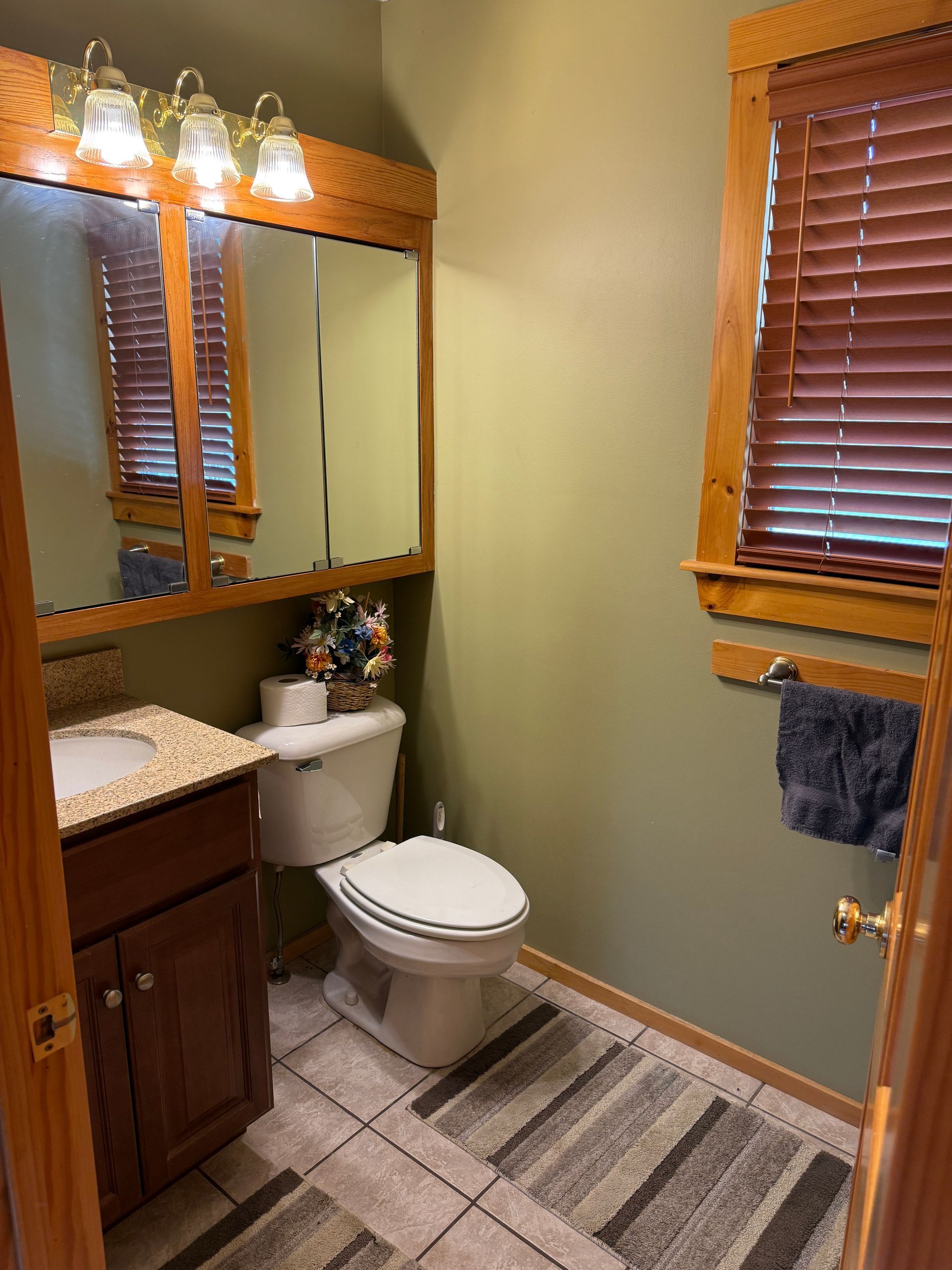Small bathroom with toilet, sink, mirror, and a window with blinds. Green walls, wood trim, and beige tile.
