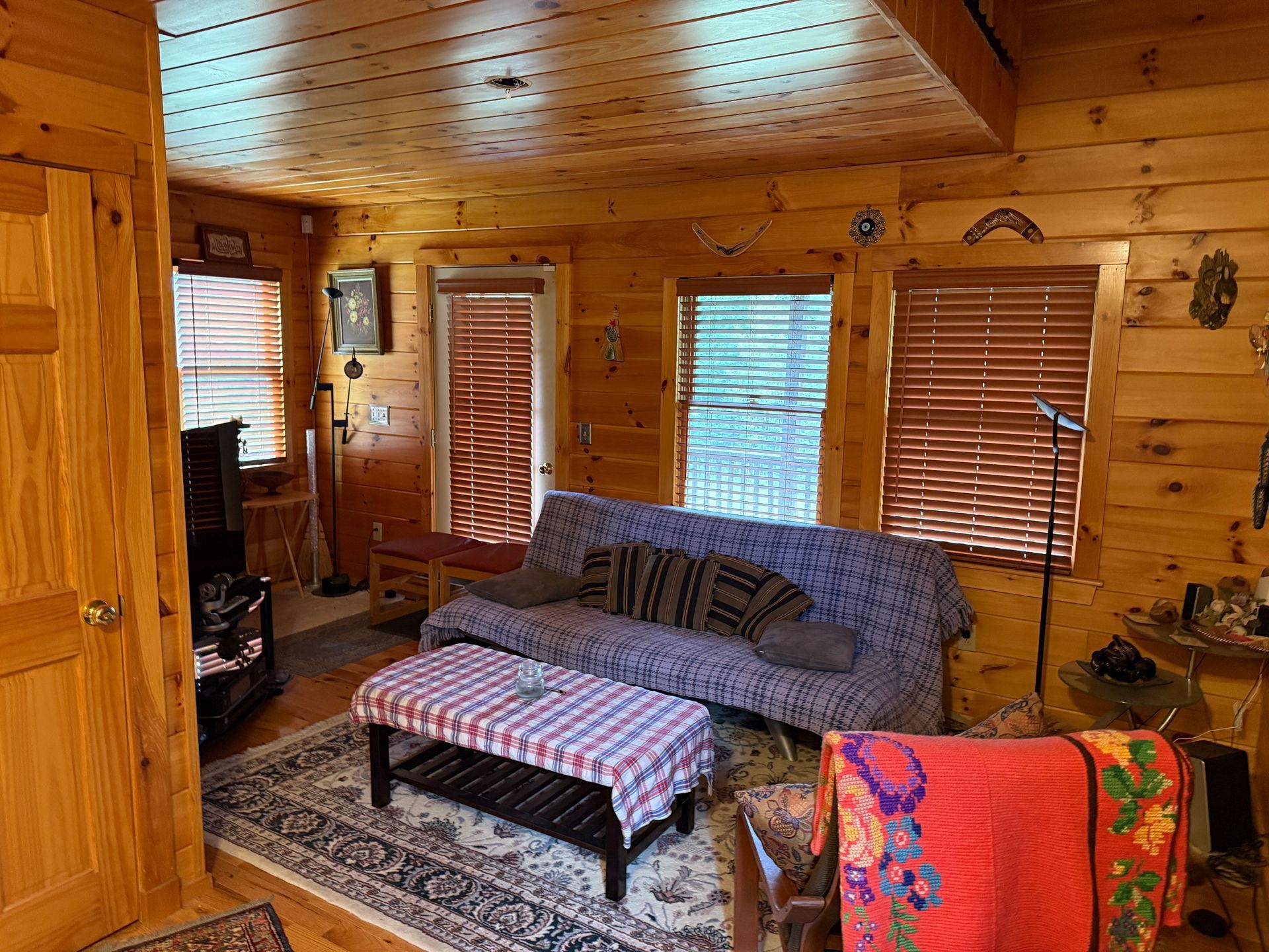 Cozy cabin interior with wood walls and ceiling, couch, coffee table, windows with blinds, and rug.
