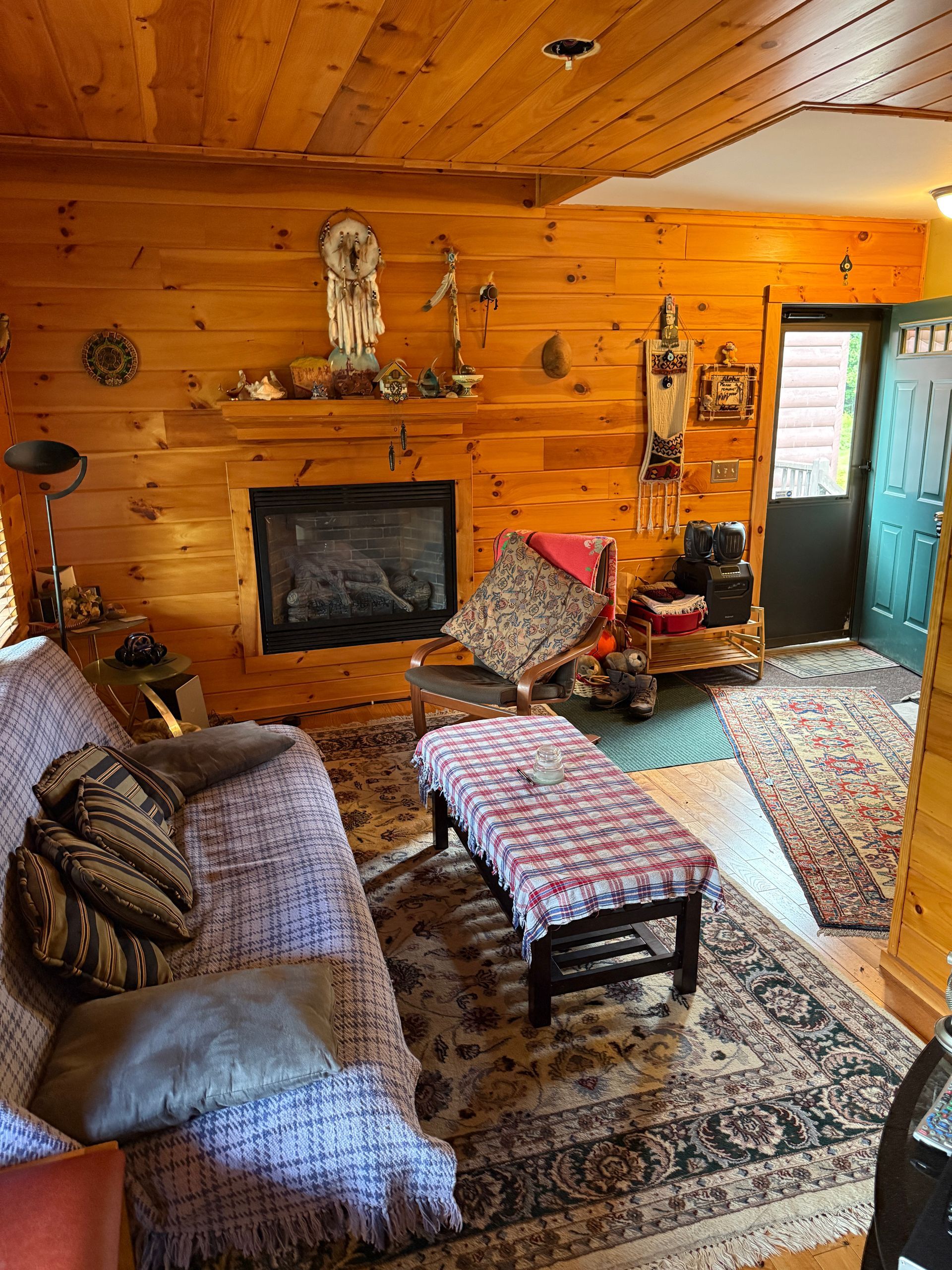 Cozy living room with a fireplace, wooden walls, a sofa, rug, and an open doorway.