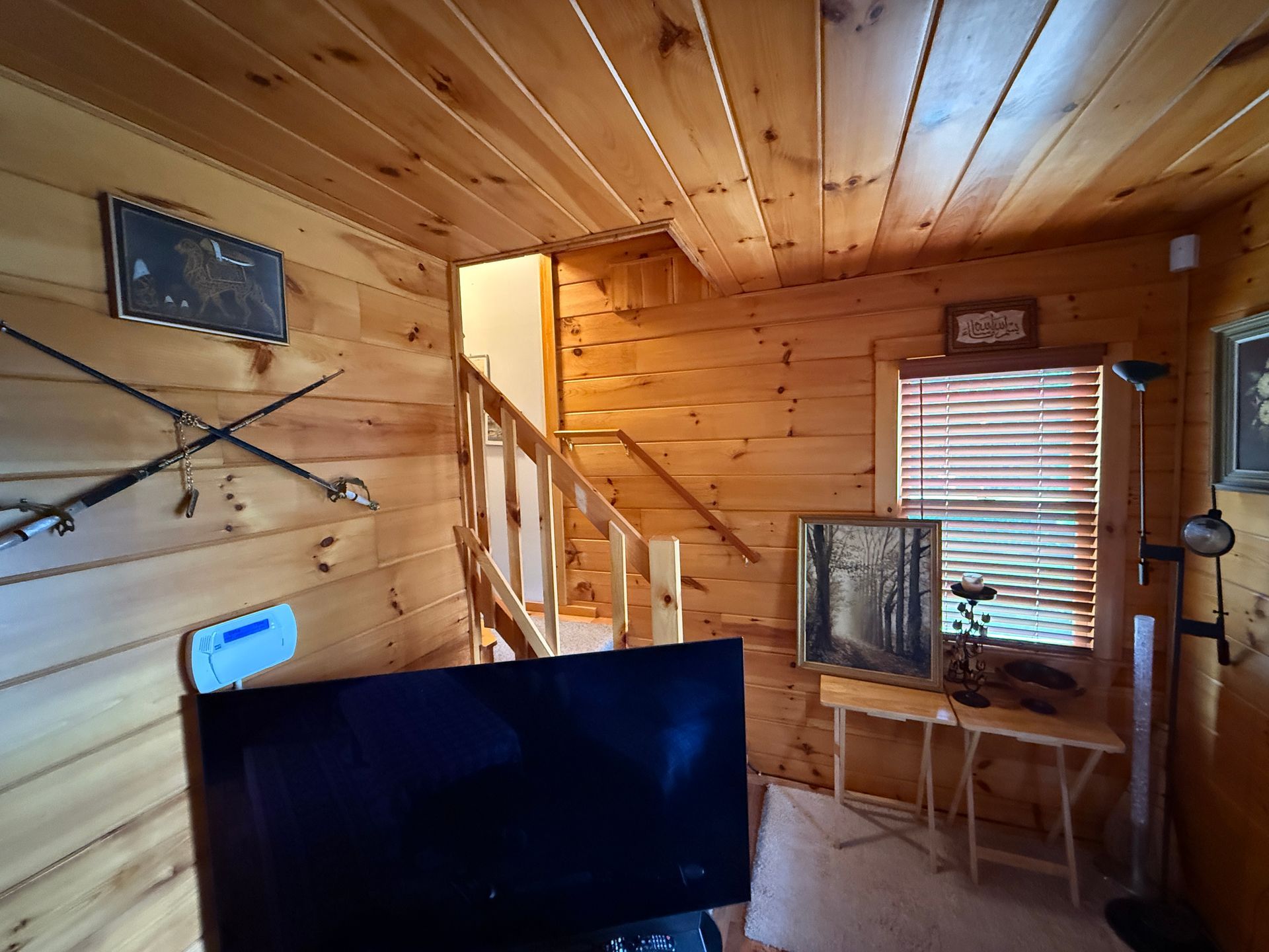 Cozy wooden cabin interior with stairs, artwork, a TV, and a window with blinds.