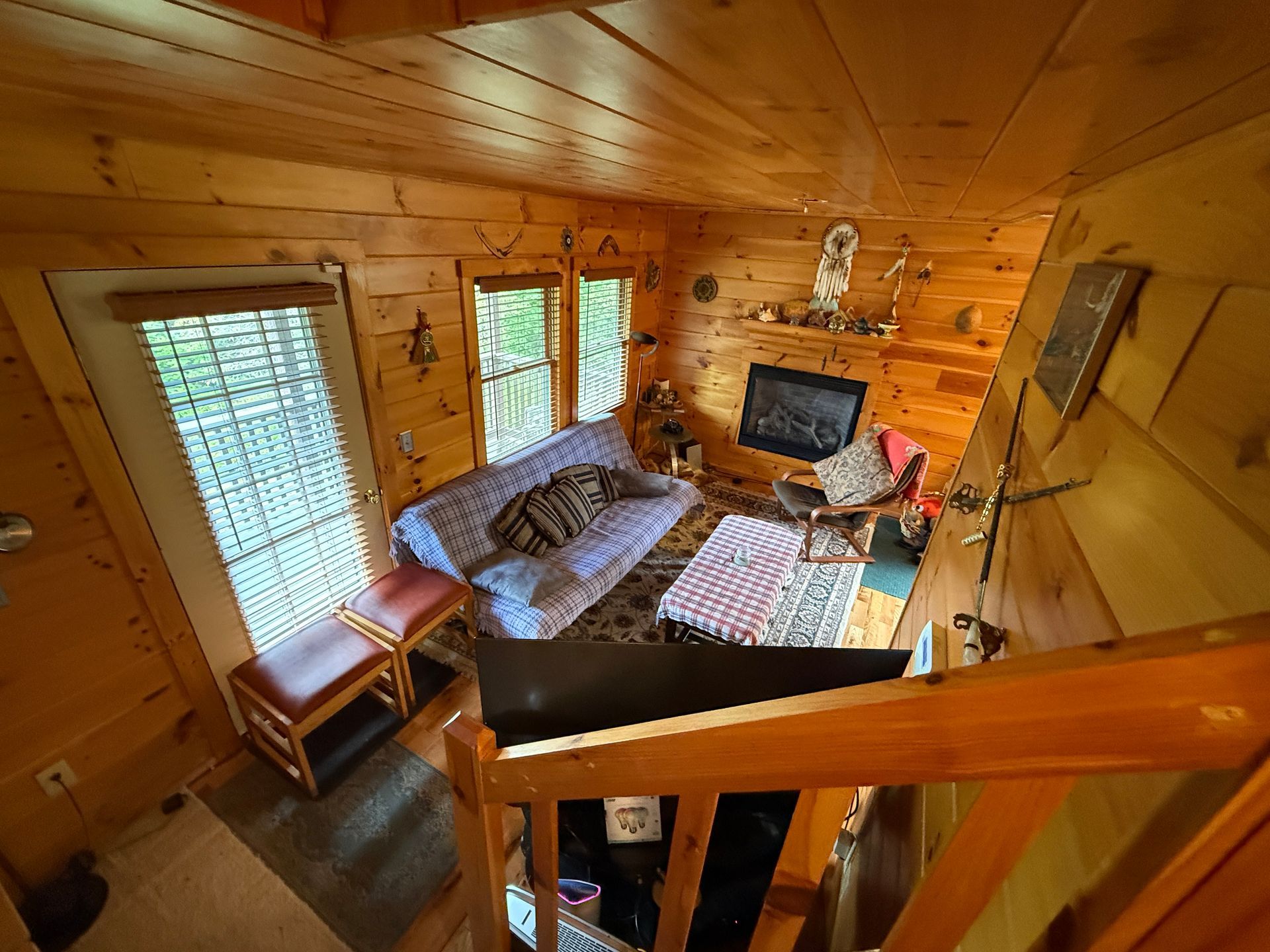 Cozy cabin interior with wooden walls, fireplace, sofa, windows, and a staircase.