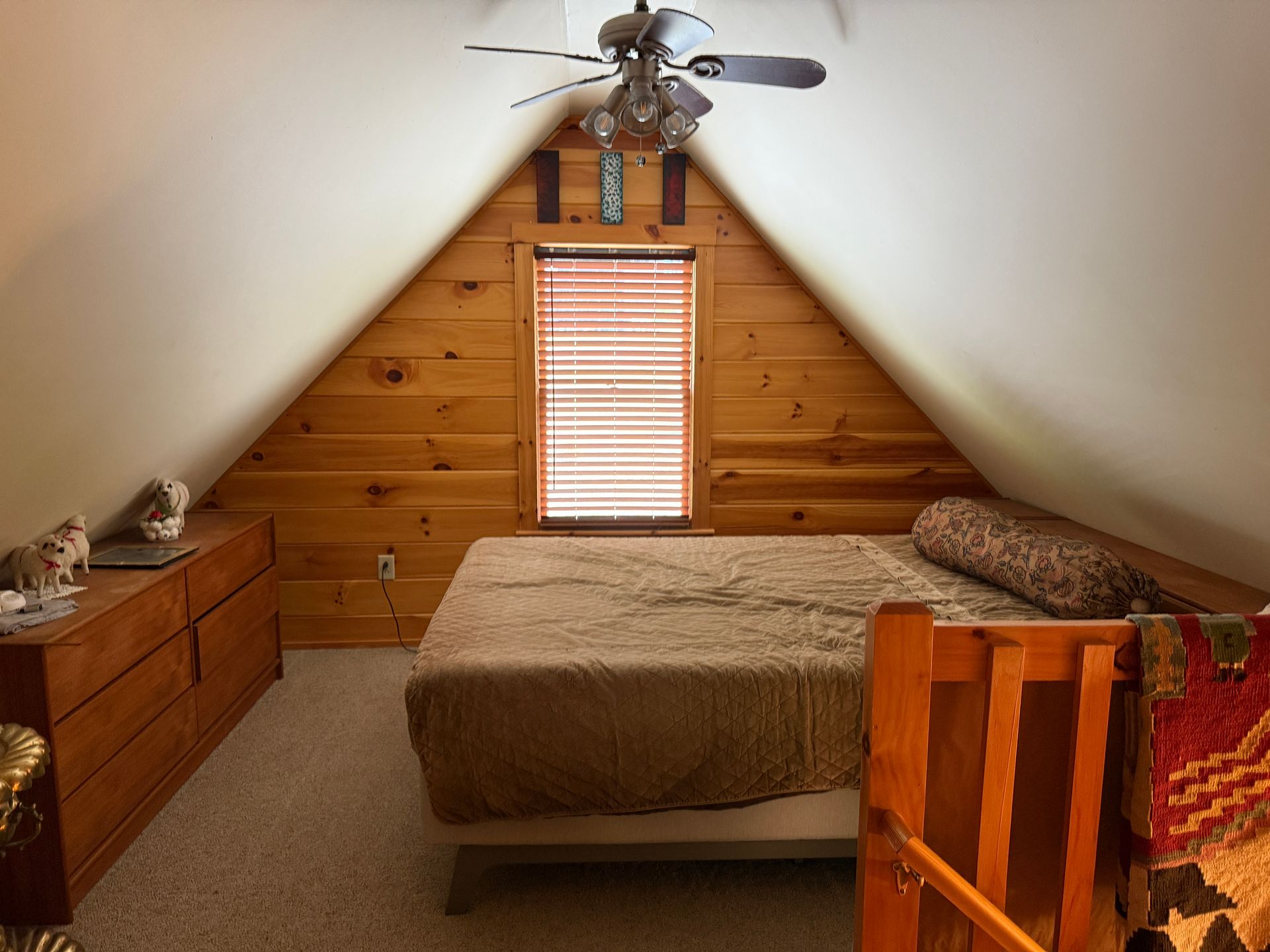 Bedroom in a cabin-style attic; wood walls, a bed, dresser, window with blinds, ceiling fan, and carpeted floor.